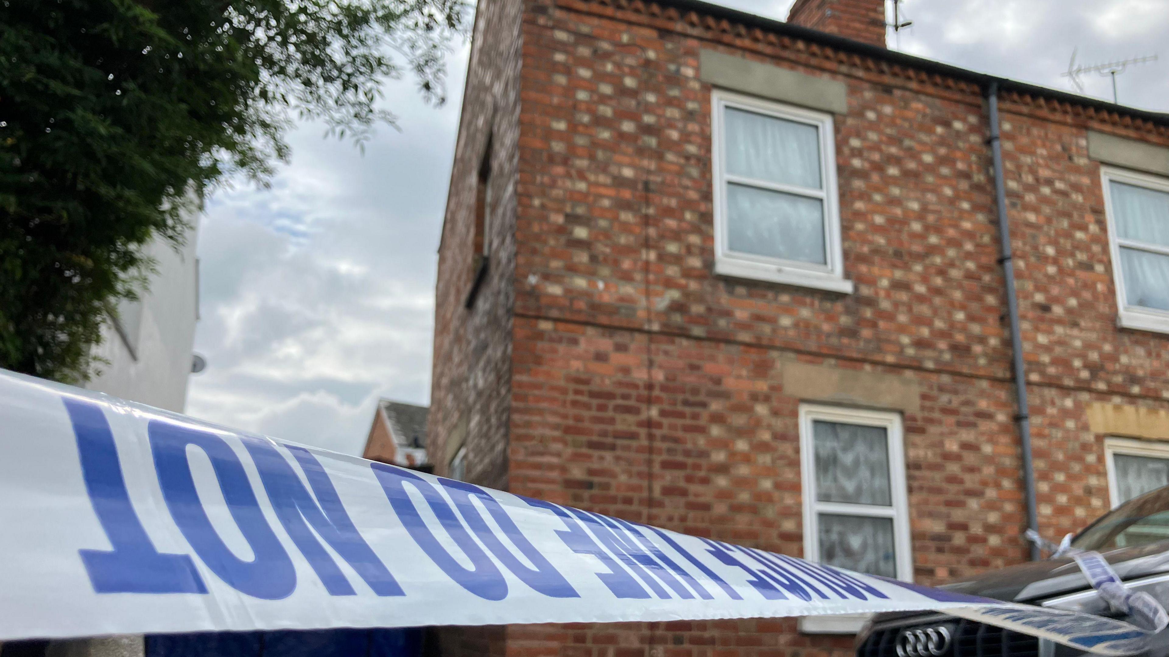 Police tape which says 'police line do not enter' in blue writing, in front of a terraced brick house with a black Audi car in front.