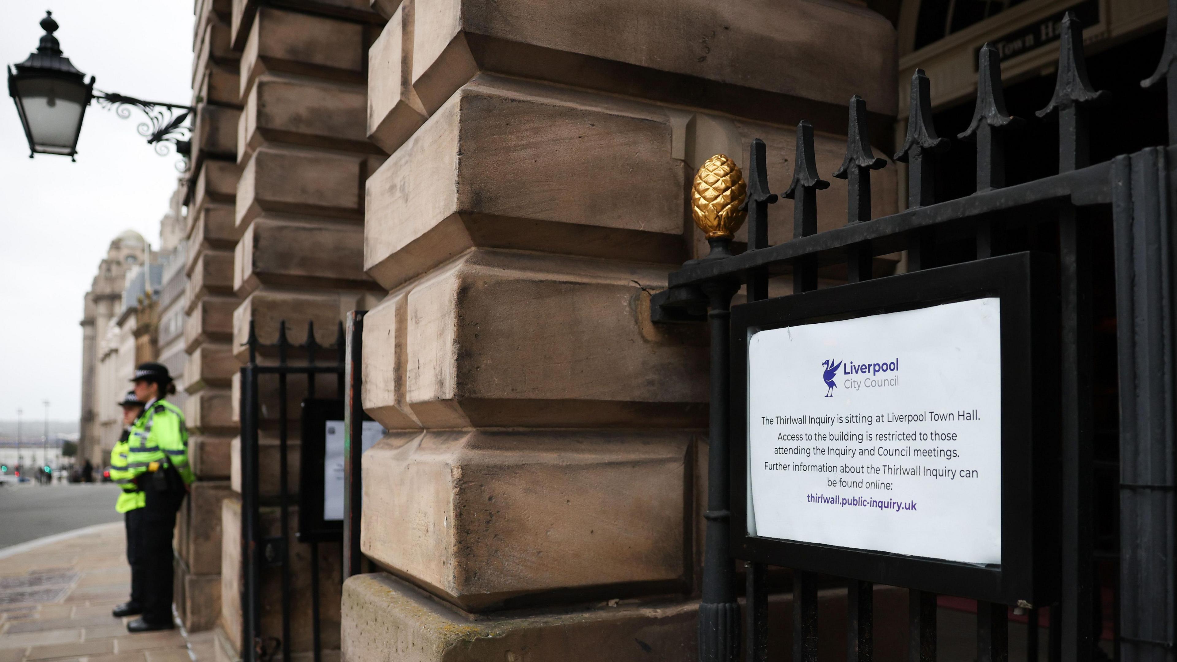 A Liverpool City Council sign with details about the inquiry's access and restrictions plus weblink for extra information is seen on a gate at the town hall, with two police officers in yellow hi-vis jackets standing guard outside the Georgian building