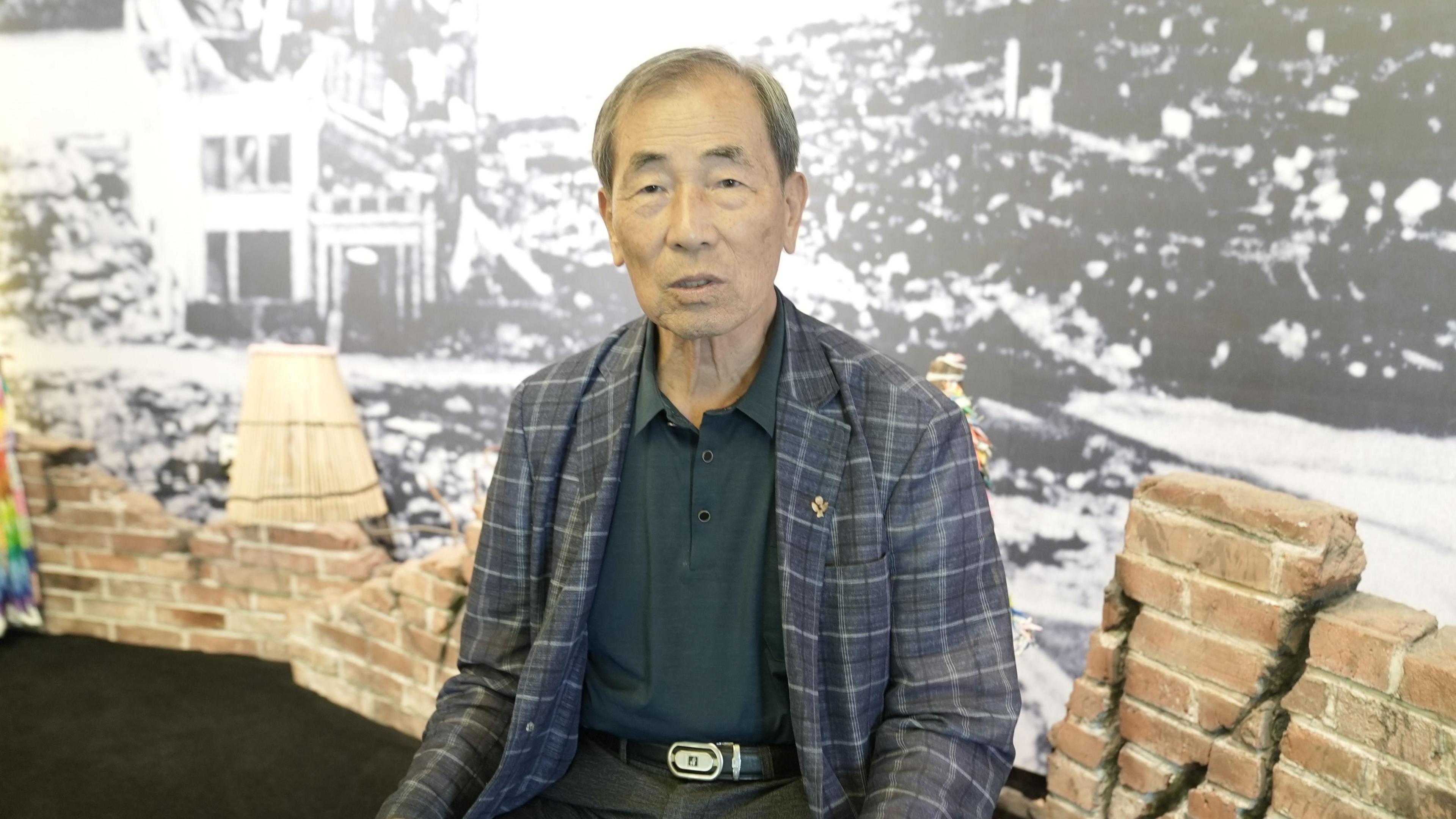 An old man in a chequered blazer sits in front of a black-and-white photograph showing destroyed streets and debris 