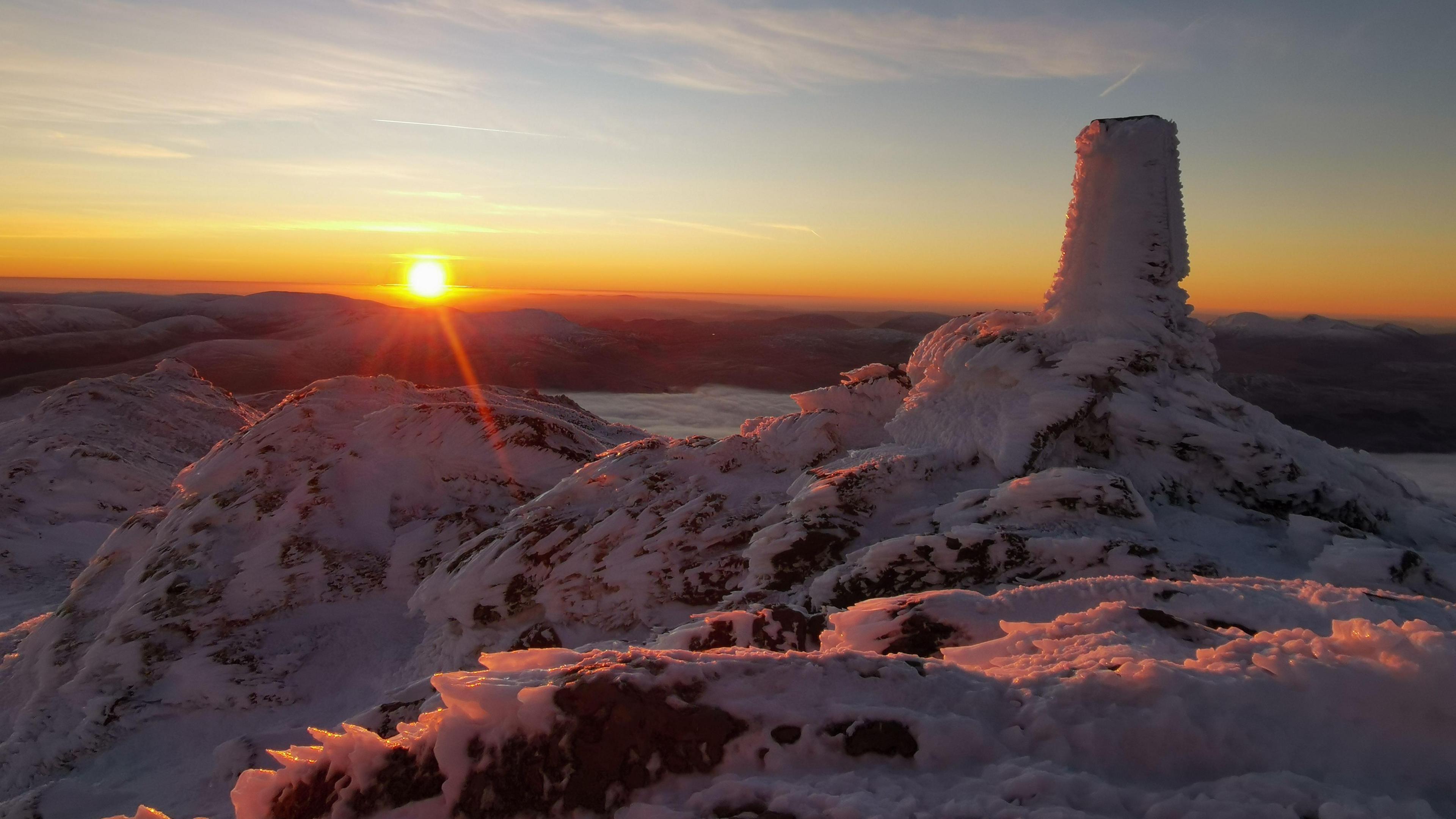 A frozen mountaintop on Ben Lawers in the Scottish Highlands. The sun is rising in the background, casting an orange glow over the image. In the foreground, large areas of the image are covered in ice and snow. The sky is light blue, fading in to orange.