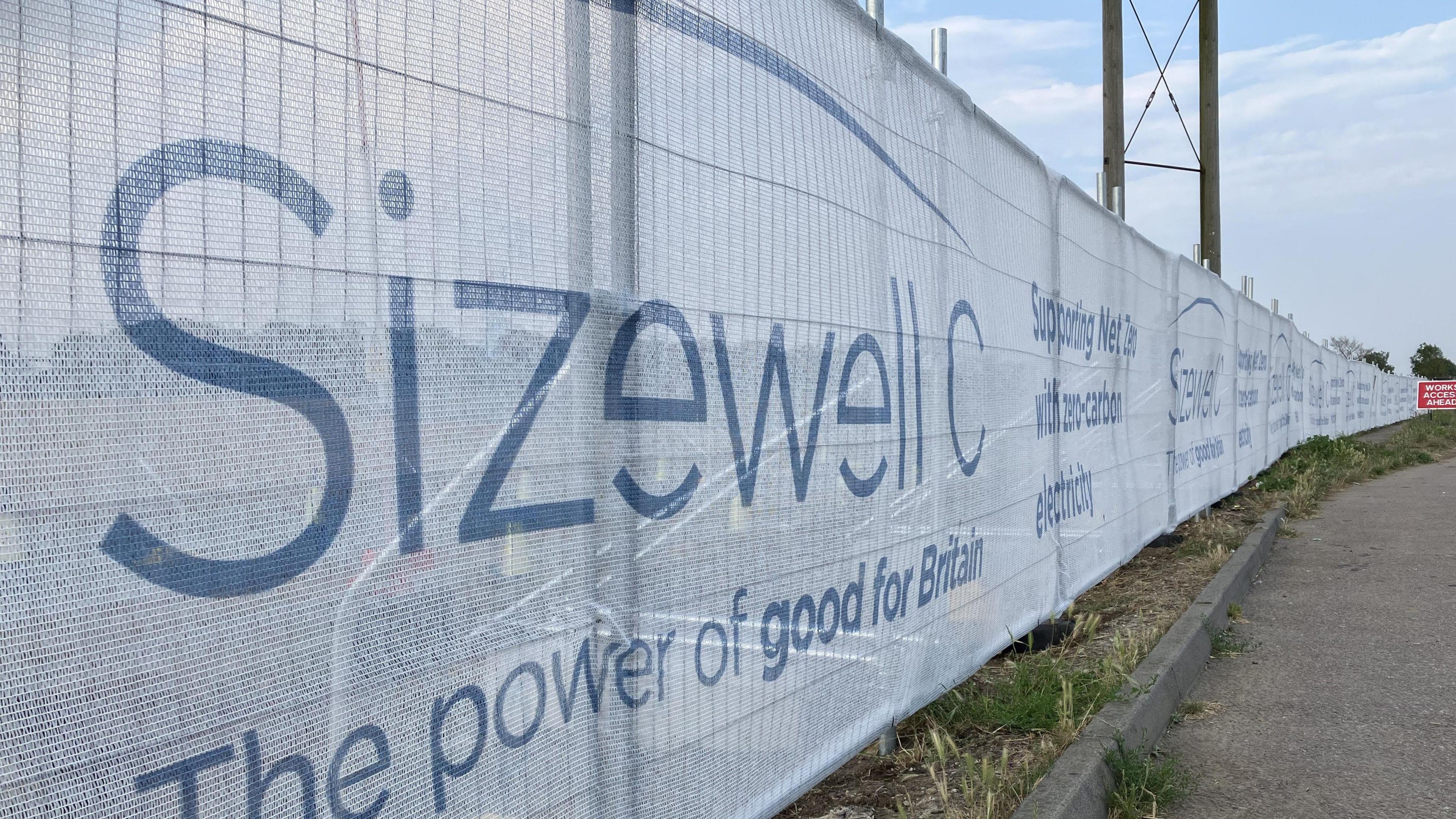 A construction fence with white fabric over it blocking off the view behind it. The words "Sizewell C, The power of good for Britain" are written in blue writing on the white fabric.