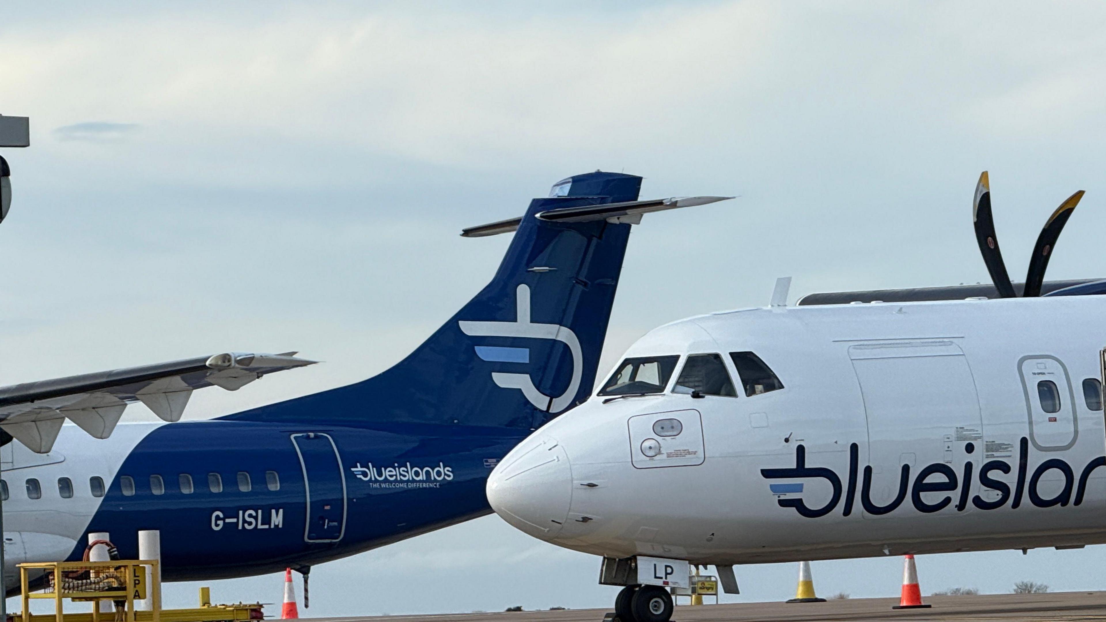 Two aircraft are stationary at Guernsey Airport. One is coloured navy blue with he logo of Blue Islands in white on the tail. The logo is an interlocking letter B and I. The second aircraft is white with the words Blue Islands in navy blue lettering.