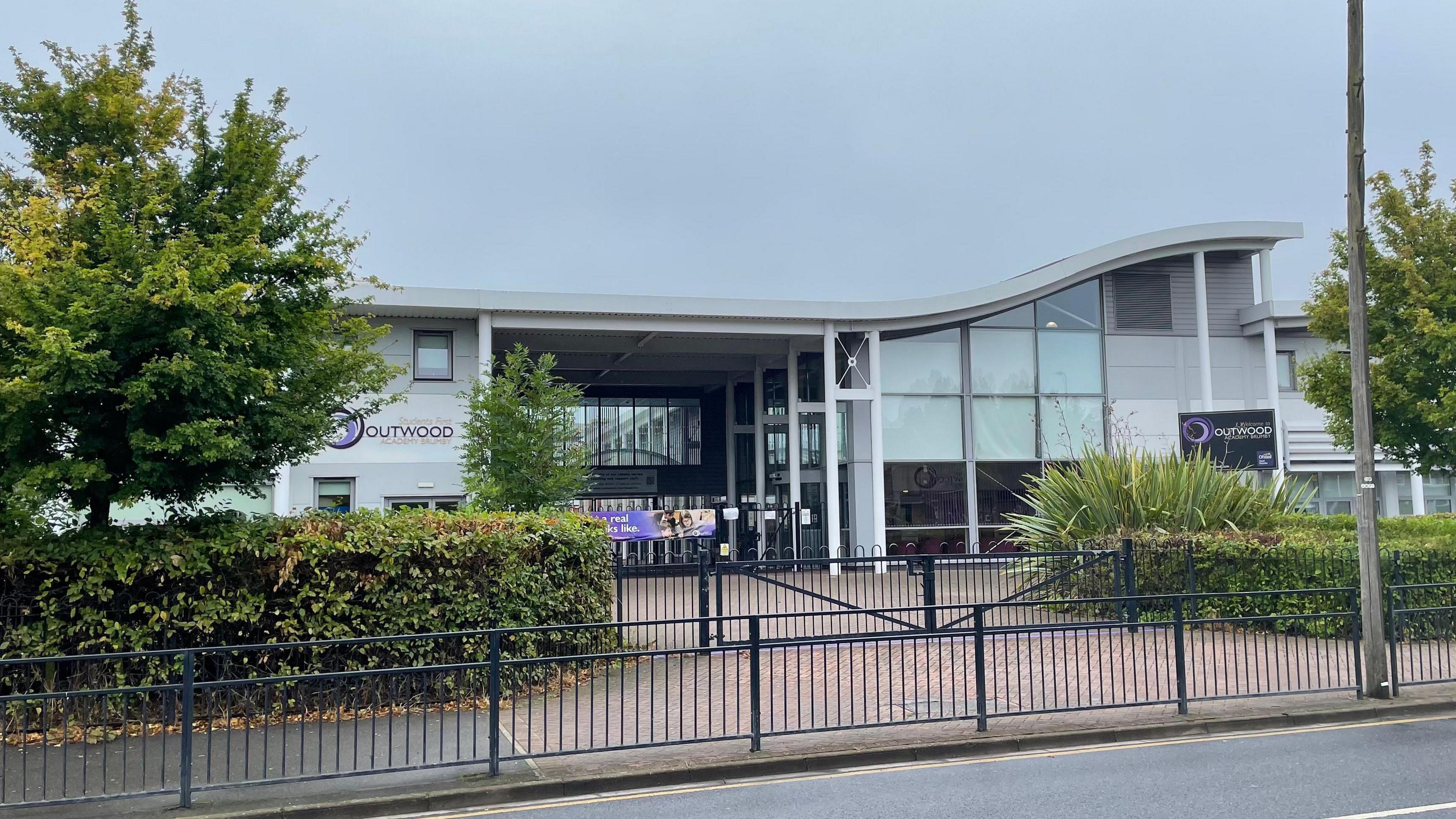 The front of the school, which has large glass windows and a curved metal frontage. There are trees and hedges at either side of the entrance.