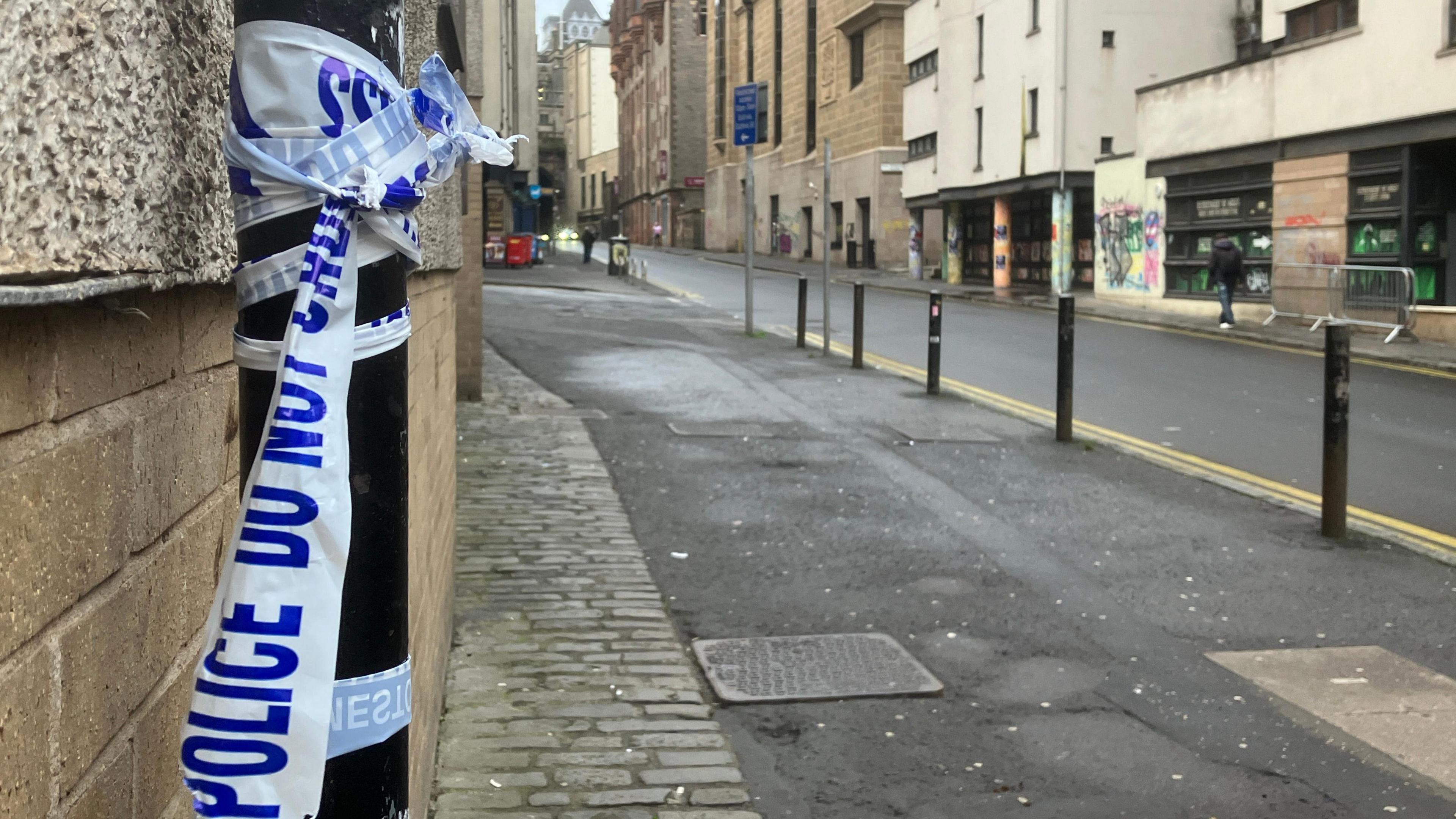 Broken police tape attached to a lamp post in the Cowgate area of Edinburgh. The tape is white, with blue writing. The street in the background is empty.