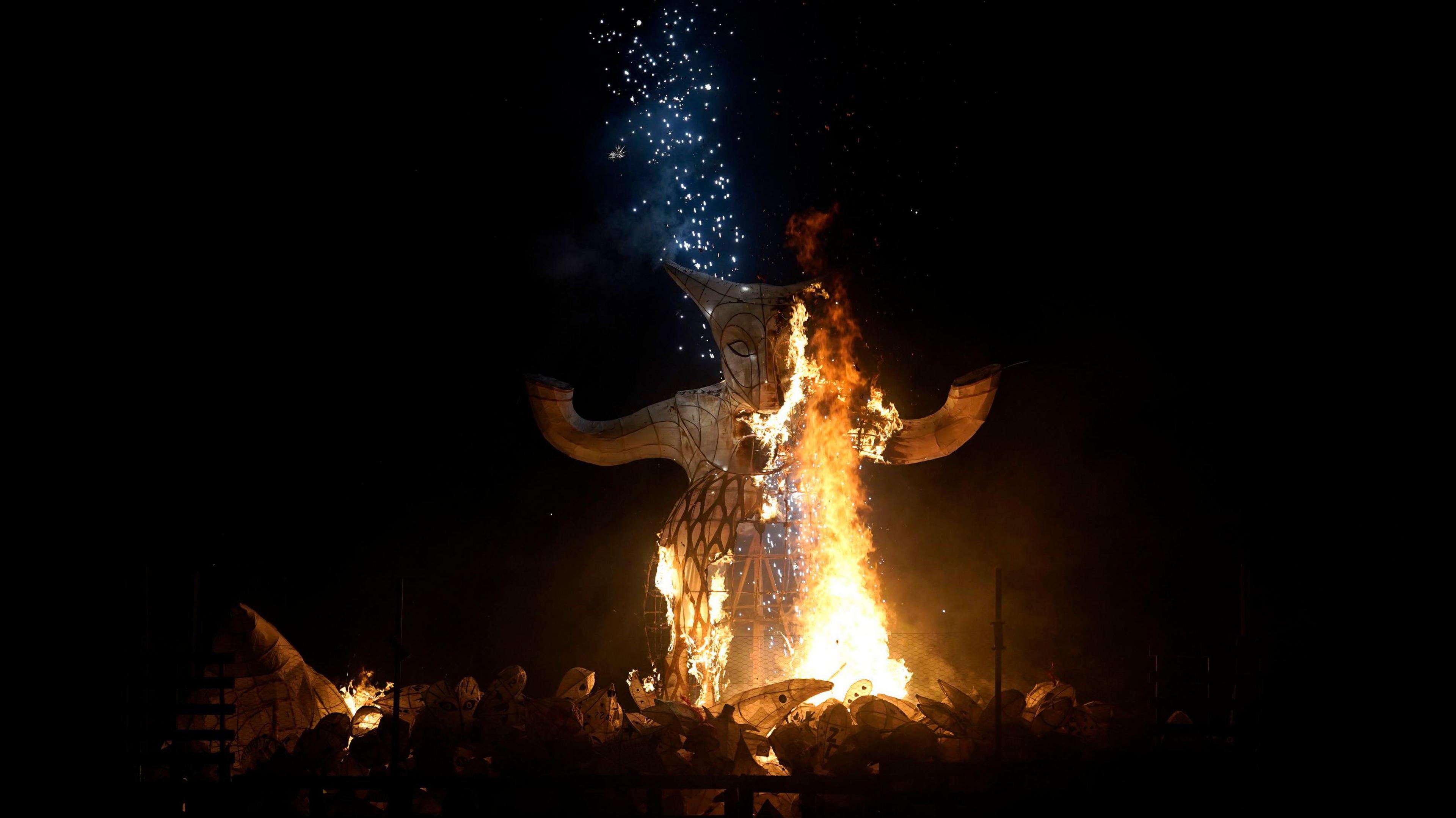 A large burning paper effigy is seen on fire, it is vaguely in the shape of a fox-like animal and there are sparks coming off the top of it.