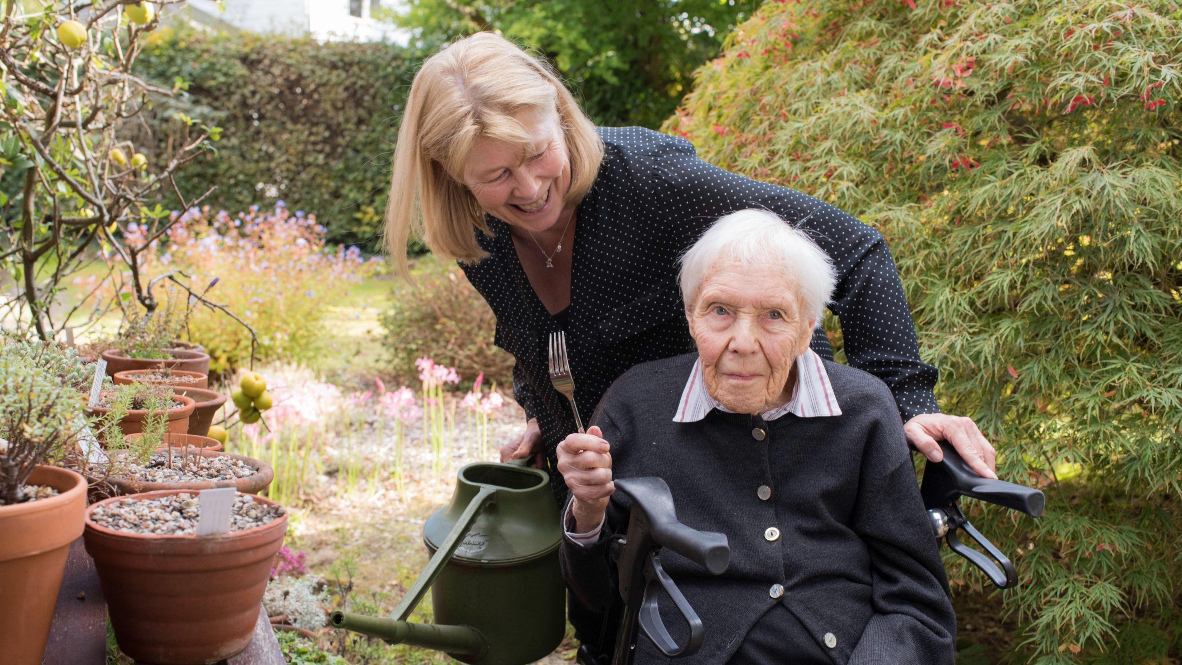 An elderly woman in a wheelchair holds up a fork. She is in a pretty garden with pots and bushes. 