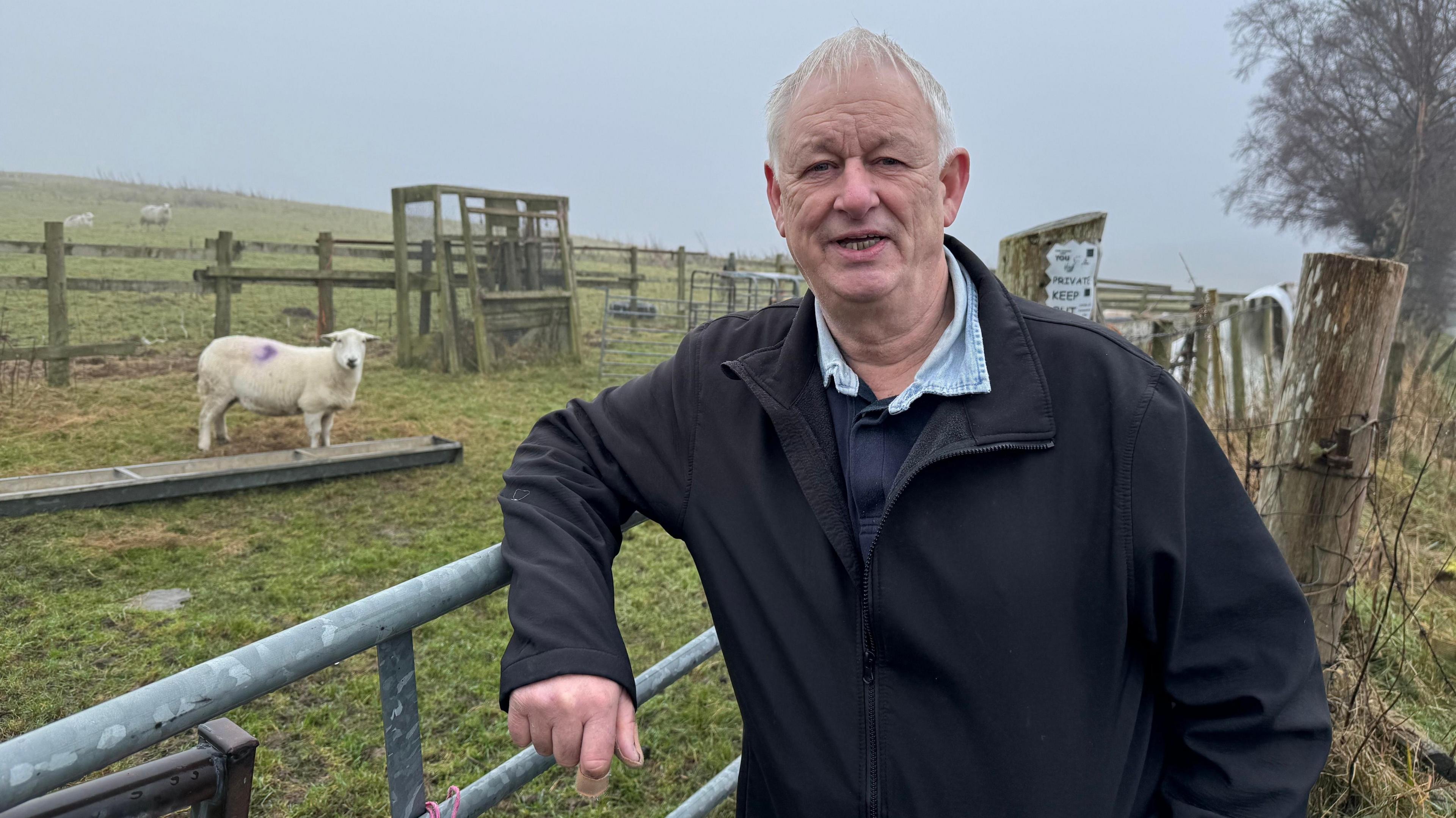 Trevor Bates stands leaning against a farm gate. He is in his sixties with white short hair, clean shaven, wearing a black jacket, a navy sweater, and a blue shirt, with the open neck collar showing. Behind him, a sheep looks on at a feeding trough in a patch of green field