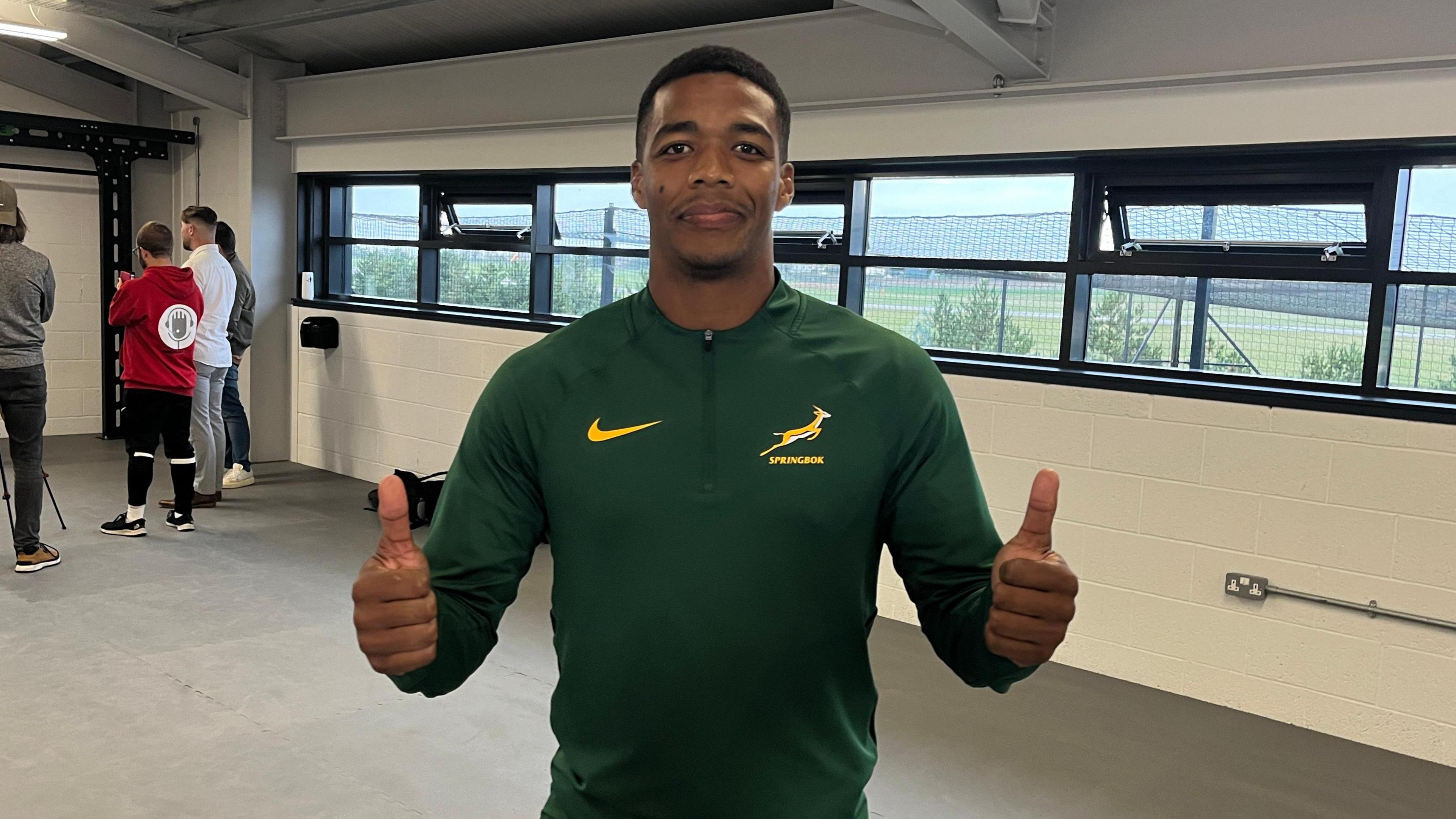 Grant Williams wearing a green Springbok T-shirt and smiling towards the camera. He has both of his thumbs up. In the background are several windows looking over the green pitch and there are people stood in the distance on the left behind him.