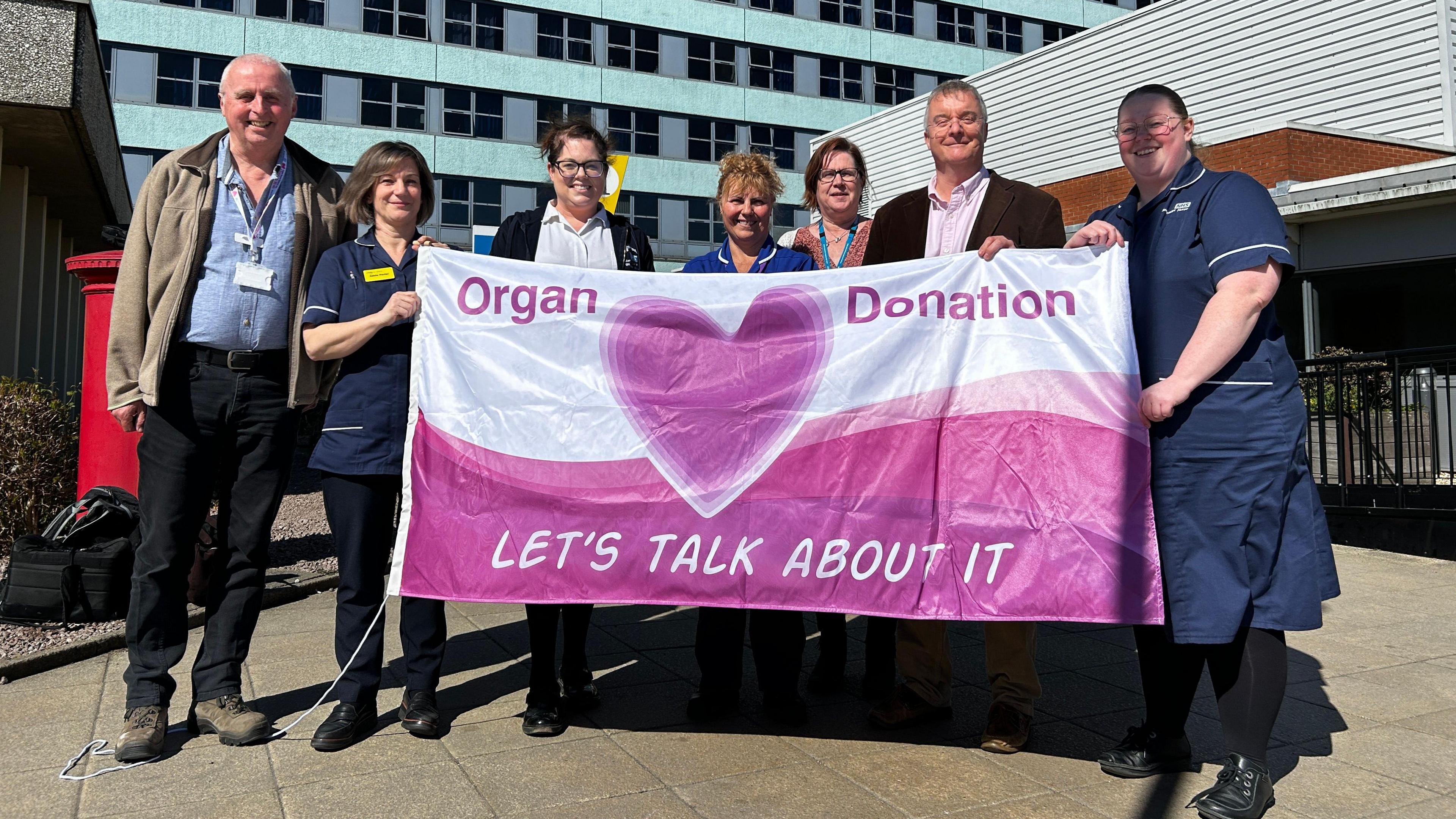 A group of two men and five women, some in uniform, holding a pink and white flag and smiling. They are standing outside a hospital. The flag has the messages "Organ Donation" and "Let's talk about it" on it, and there is a pink heart in the middle.