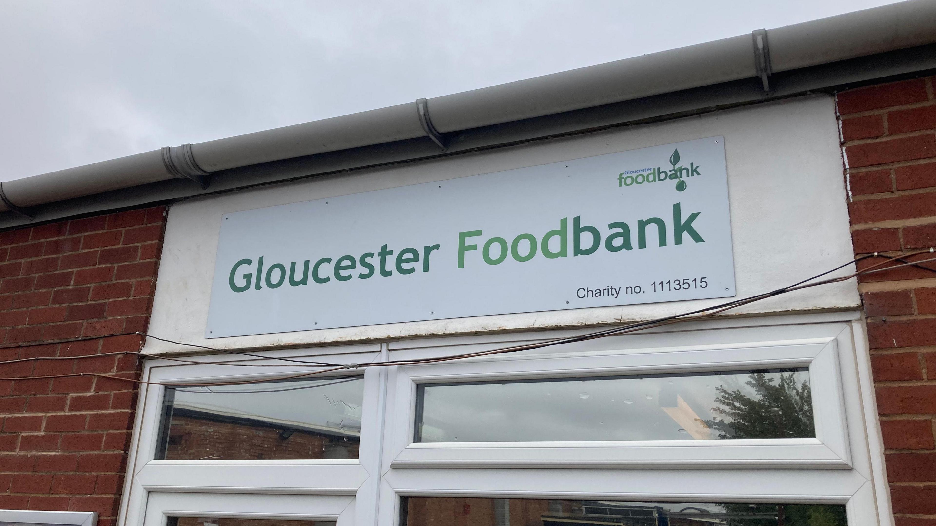A white plastic sign with Gloucester Foodbank in green letters is stuck to the brick wall above the entrance door.