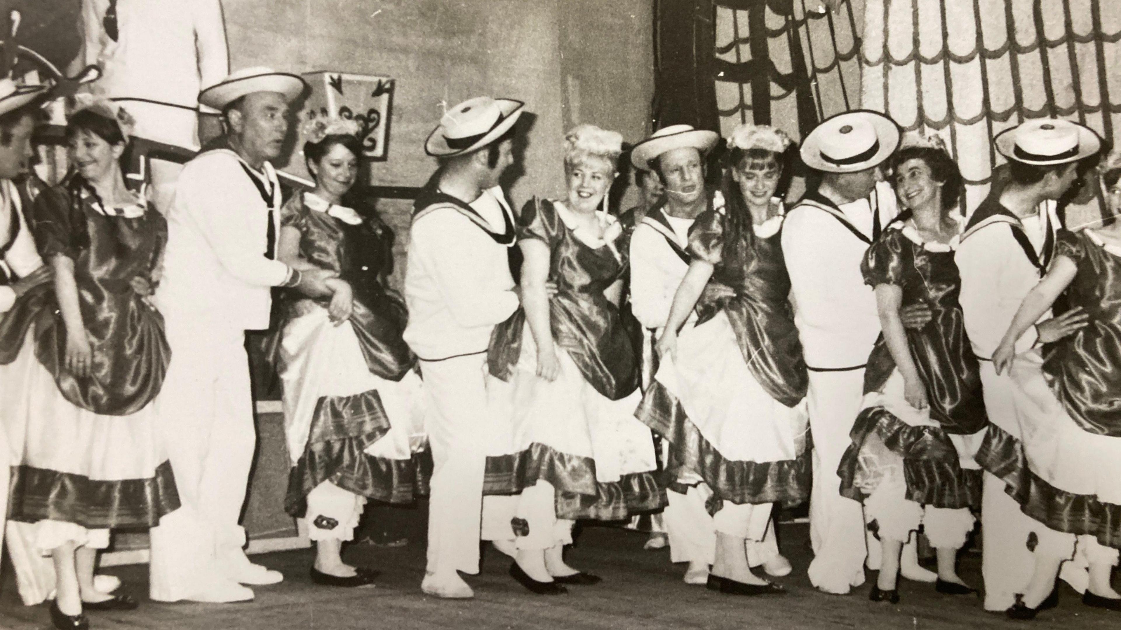 A black and white image showing men and women on stage. The men are in light colour sailor-type costumes and the women are wearing darker coloured dresses. Their costumes have a naval theme.