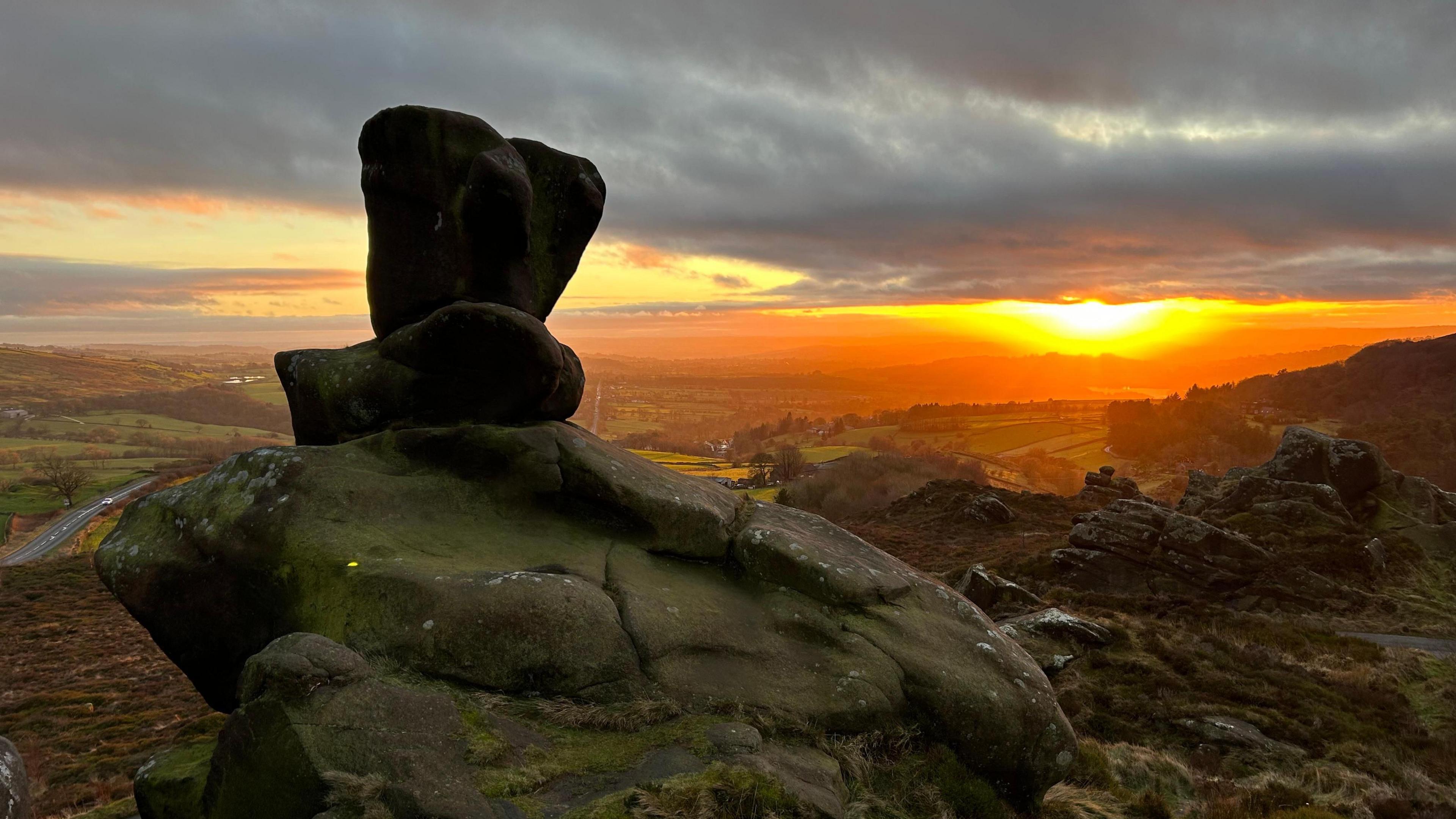 A rock stands on top of another rock on top of a hill. Behind are other hills rolling away into the distance. The sun can be seen on the horizon, peeping between clouds and lighting them up in orange and yellow.