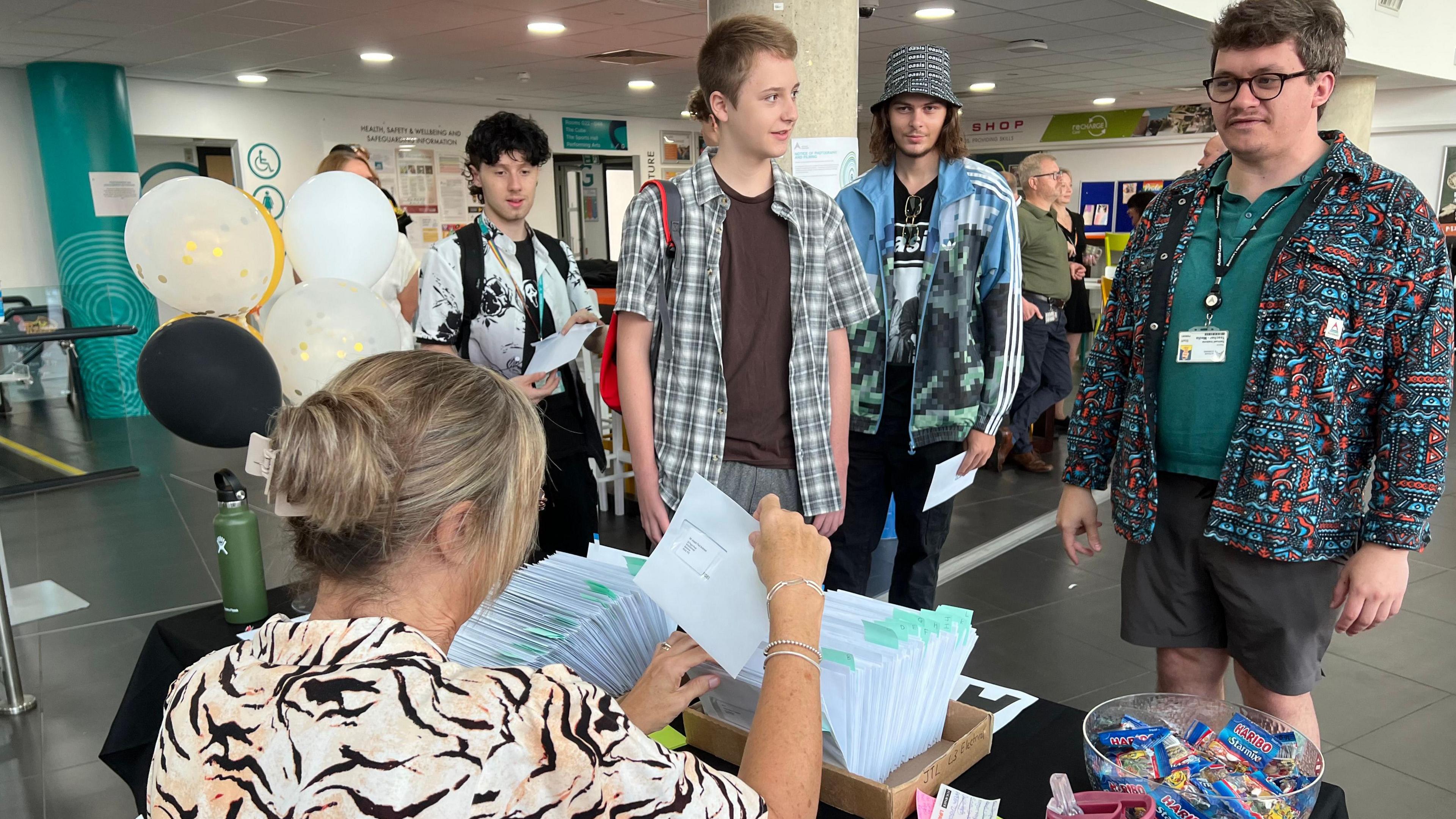 A group of four male students are waiting in front of the desk where results are being handed out. They are wearing casual clothes and standing in the college hallway. There is a woman with blonde hair pulled back into an up do and wearing a tiger print blouse, she is facing away from the camera and going through boxes filles with pieces of paper.