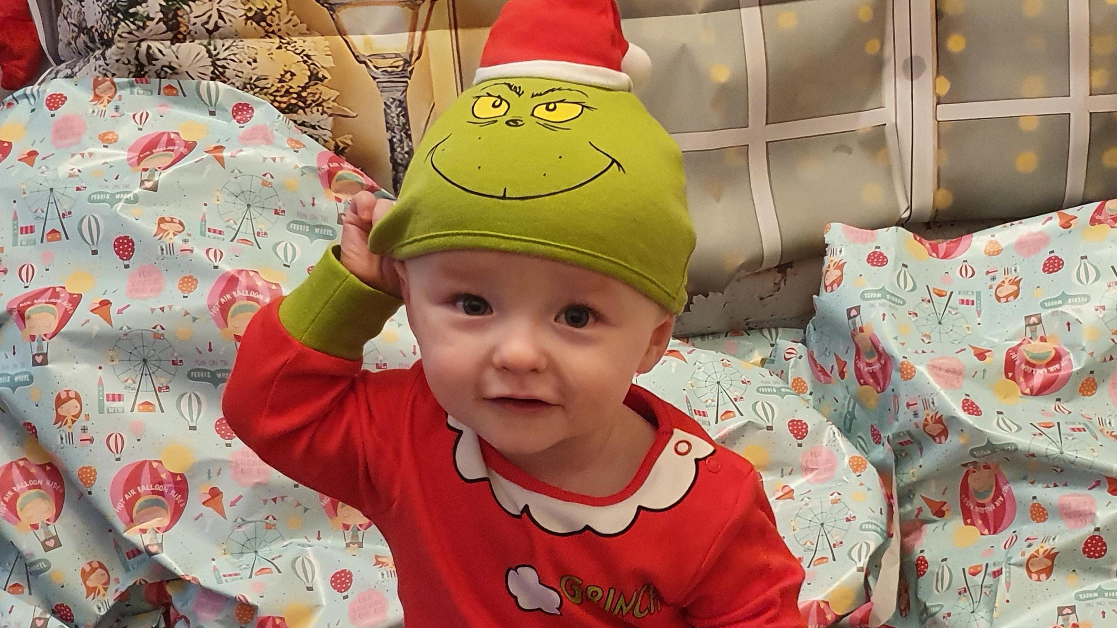 Melanie Wallwork's son Jake puts his hand against his head while smiling wearing a Christmas-themed Grinch at and onesie in front of a pile of presents. 