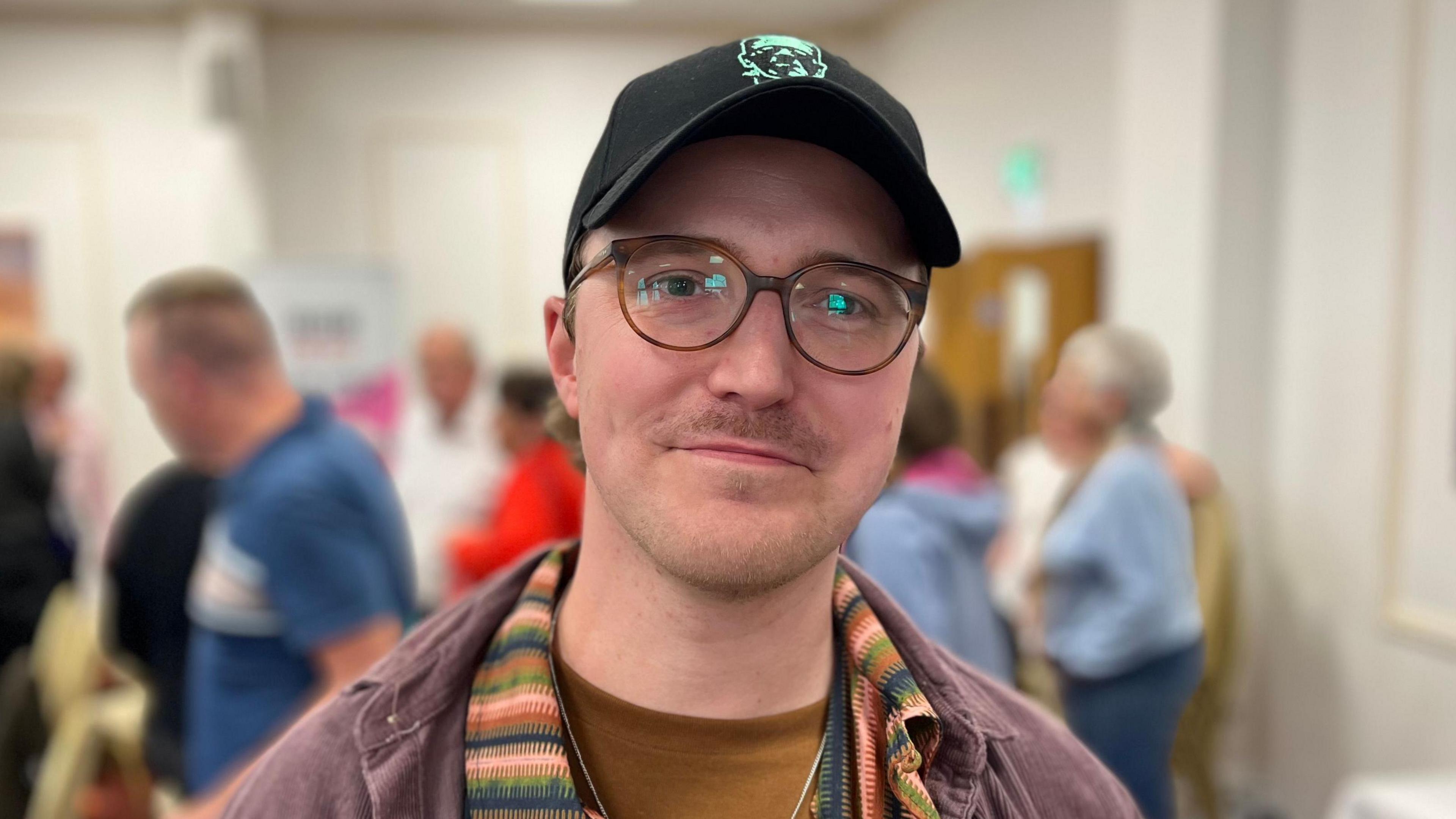 A man with a black peaked cap, wearing blue glasses. He has a small moustache.