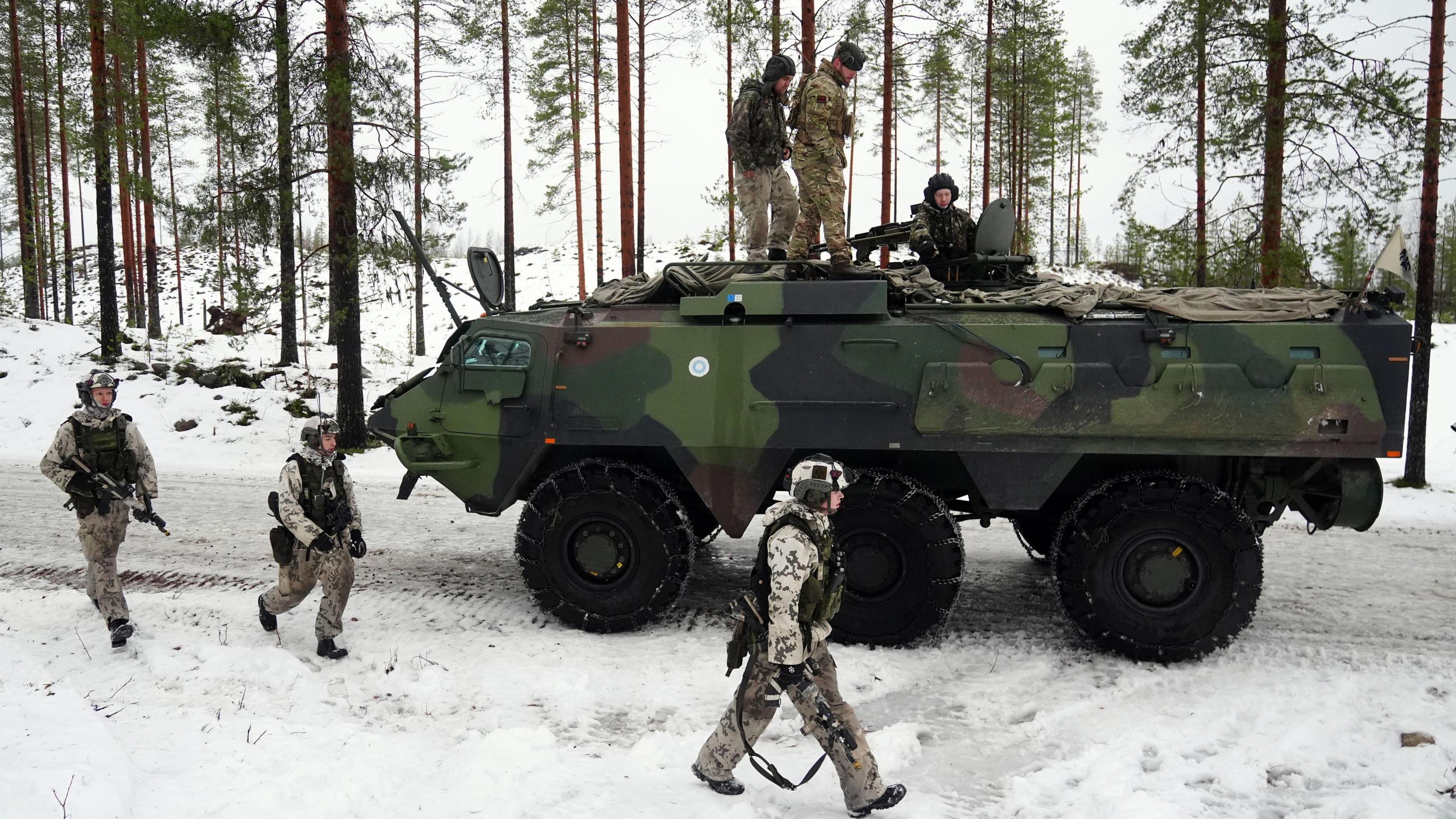 An armoured vehicle on a snow-covered road. There are three soldiers on top of the tank and three others walking along side it. 
