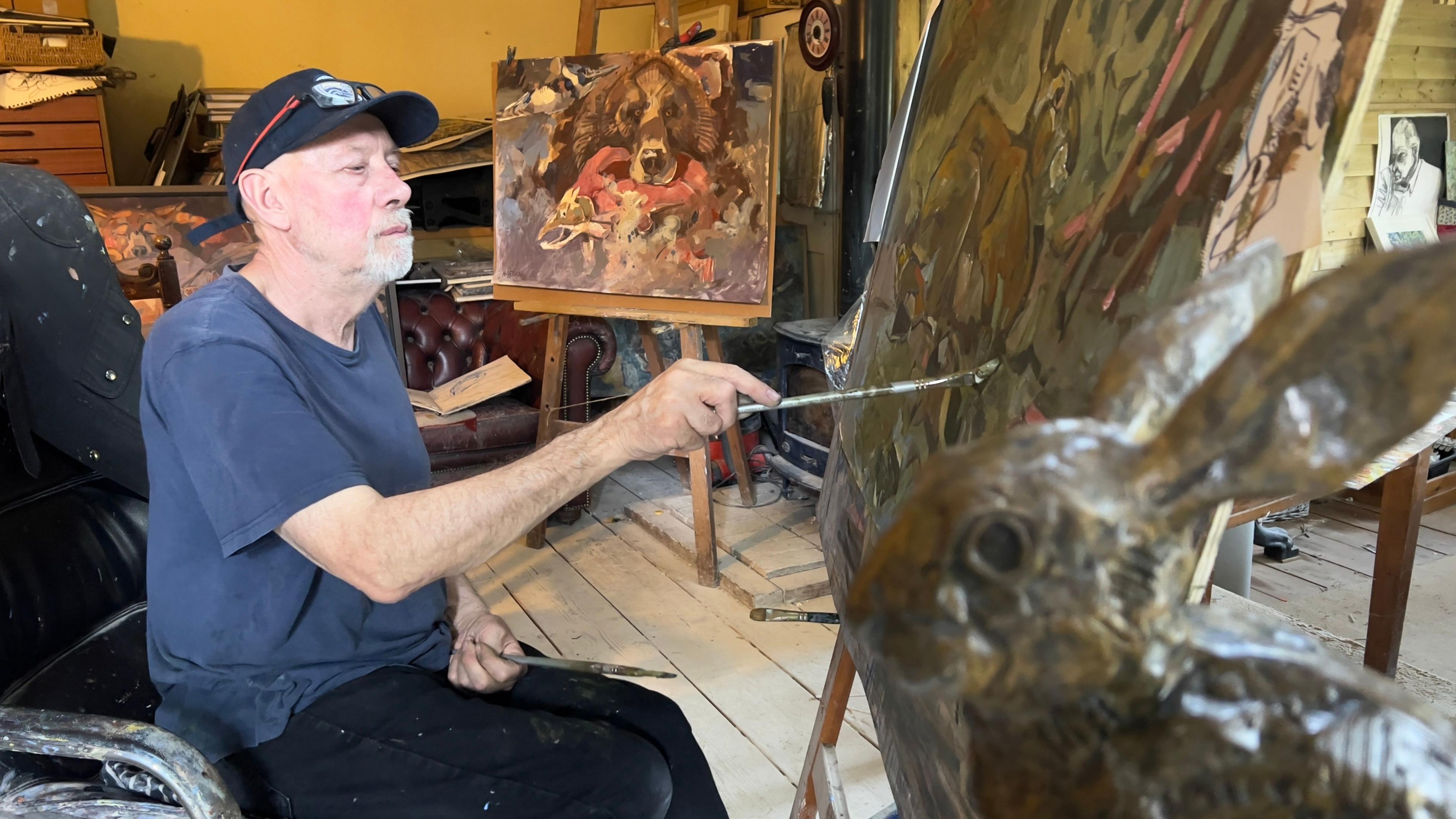 Artist Andrew Haslen is pictured in his art studio painting on a canvas. He is sitting down while wearing a blue t-shirt, black trousers and a black cap with his glasses resting on it. A sculpture of a hare sits near him while other pieces of his work surround him.