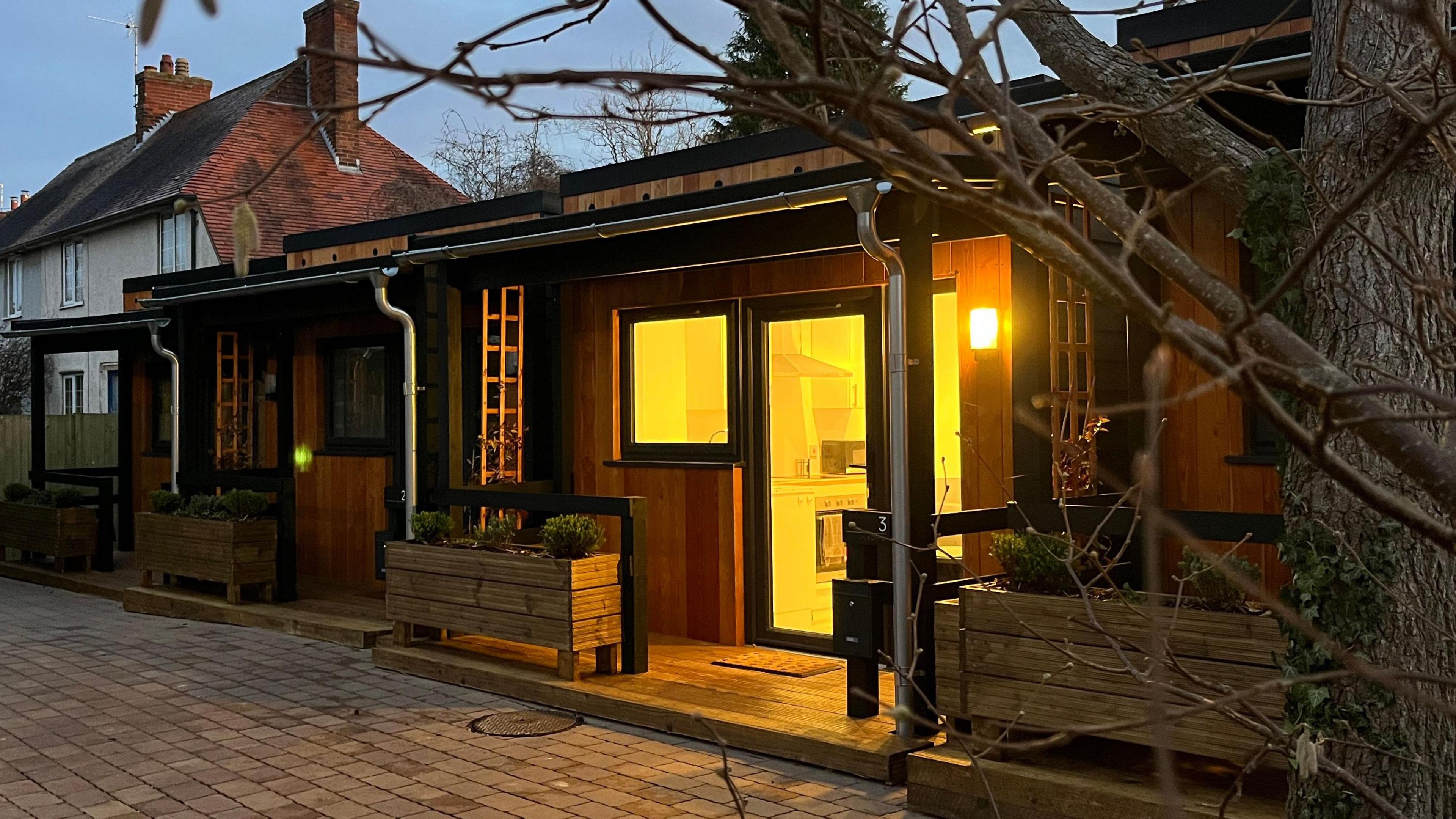 A rectangular one floor wooden building with three modular homes. Each home has a black window and front door inside an open porch area. They are separated by flower beds and trellis. The home closest to the camera has its orange lights on. It is dark outside and the light shows a kitchen inside the house with a cooker, counters and a sink.