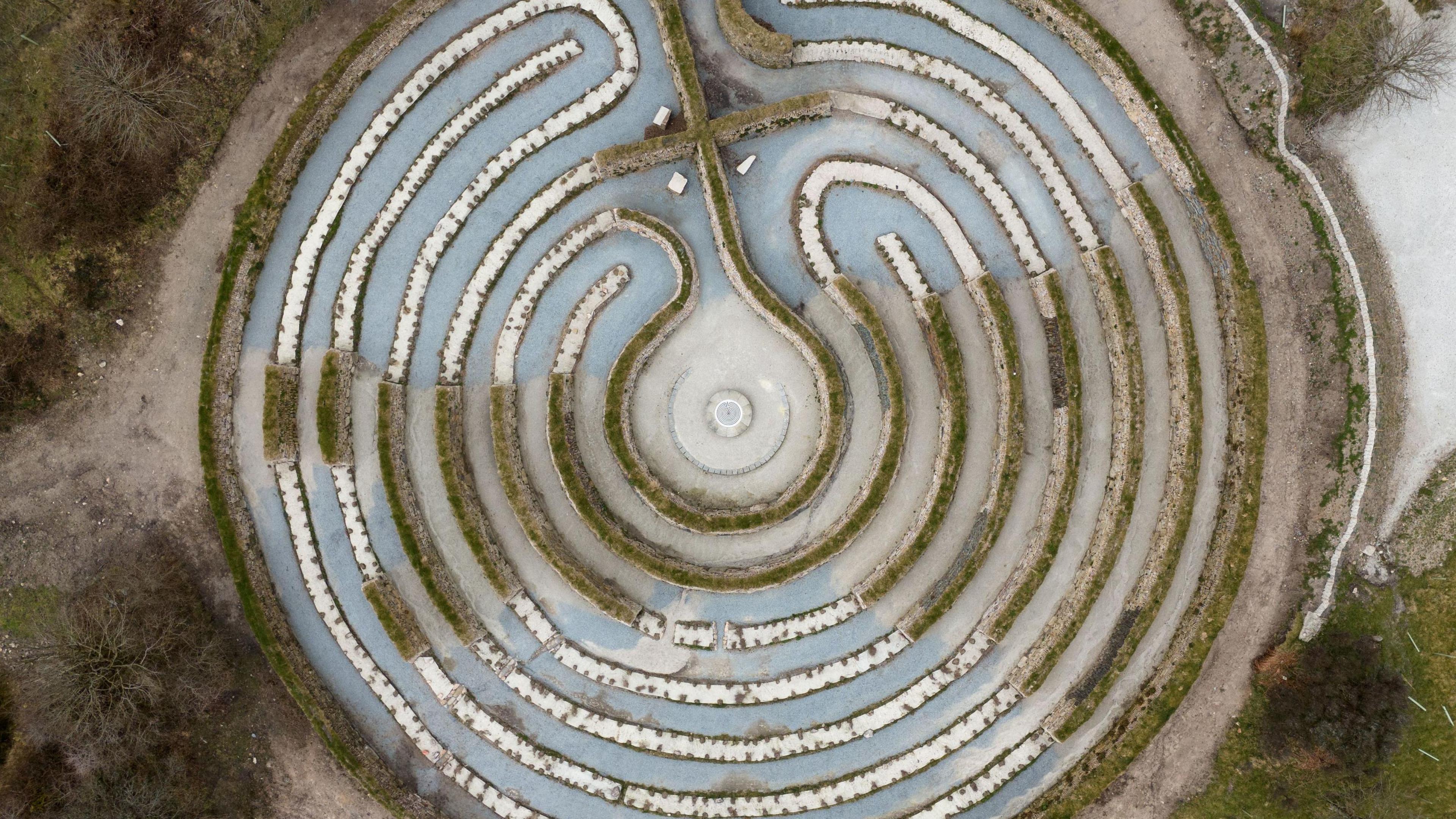 The picture shows a drone aerial shot looking straight down at a circular labyrinth. There are lots of paths separated by low hedges and walls. At the centre there is a small circular area. There is lots of greenery in the surrounding area.