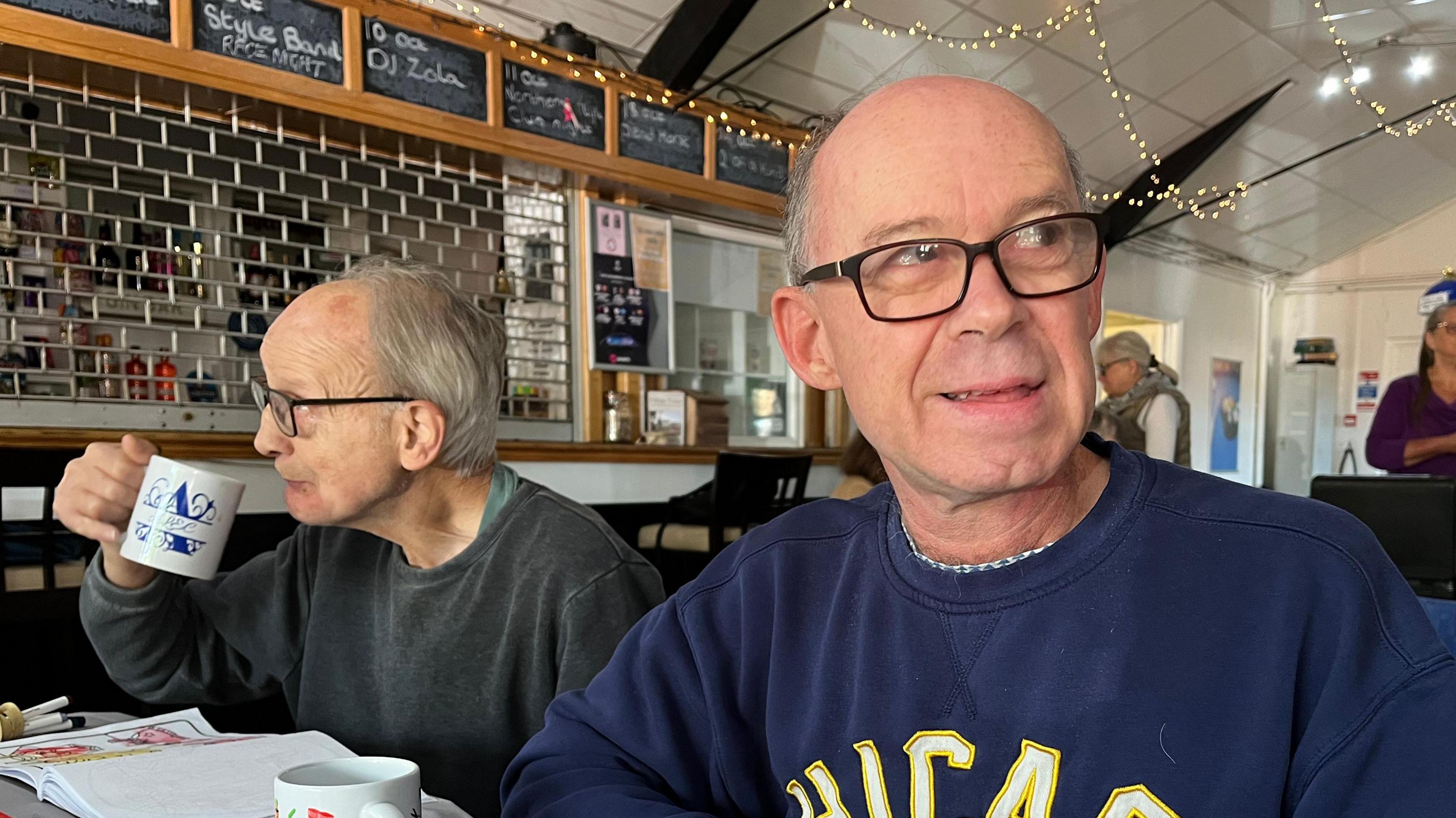 Mark is one of those who attends the weekly phobbies centre. He is wearing a blue sweatshirt, wears black glasses and sits with a cup of tea in front of him. 