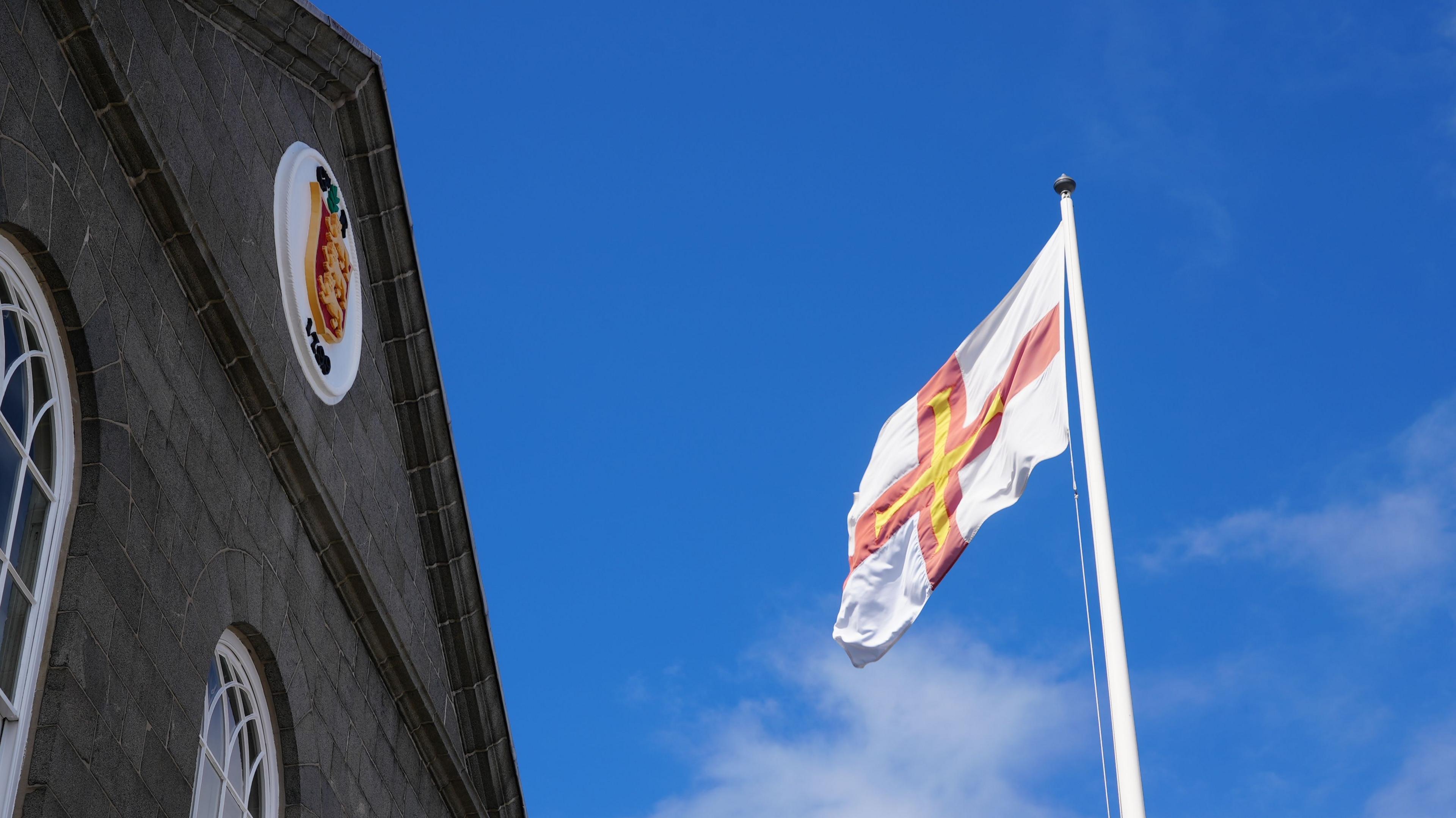 A Guernsey flag flying next to a grey granite building with a States of Guernsey crest at the top in a white circle against the background of a bright blue sky with some whispy white clouds in it