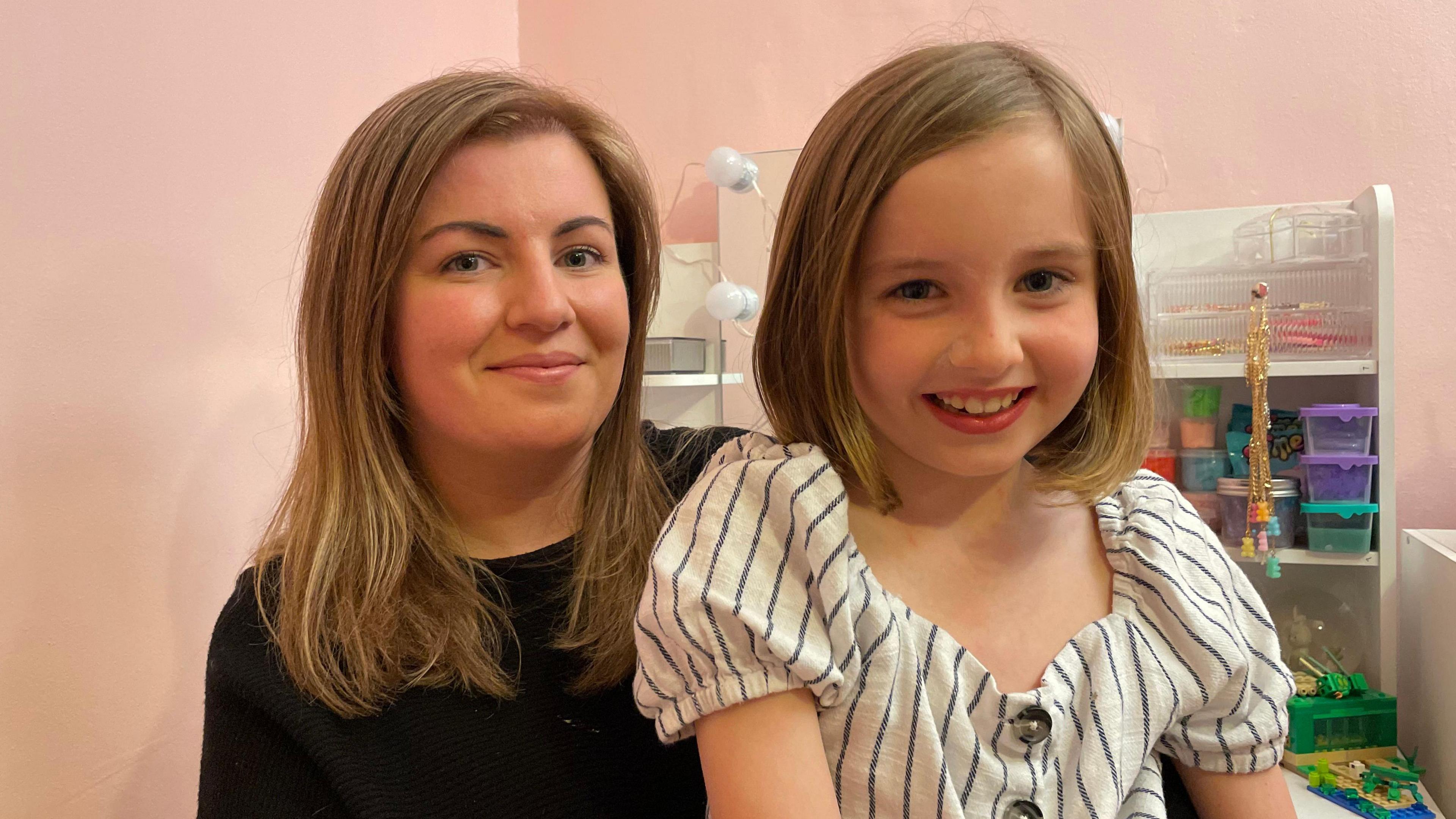 Mother and daughter Gemma and Alice. Both have dark blonde hair and are smiling. The mother is wearing a black top and her daughter is wearing a stripy white and navy top with buttons down the front. 