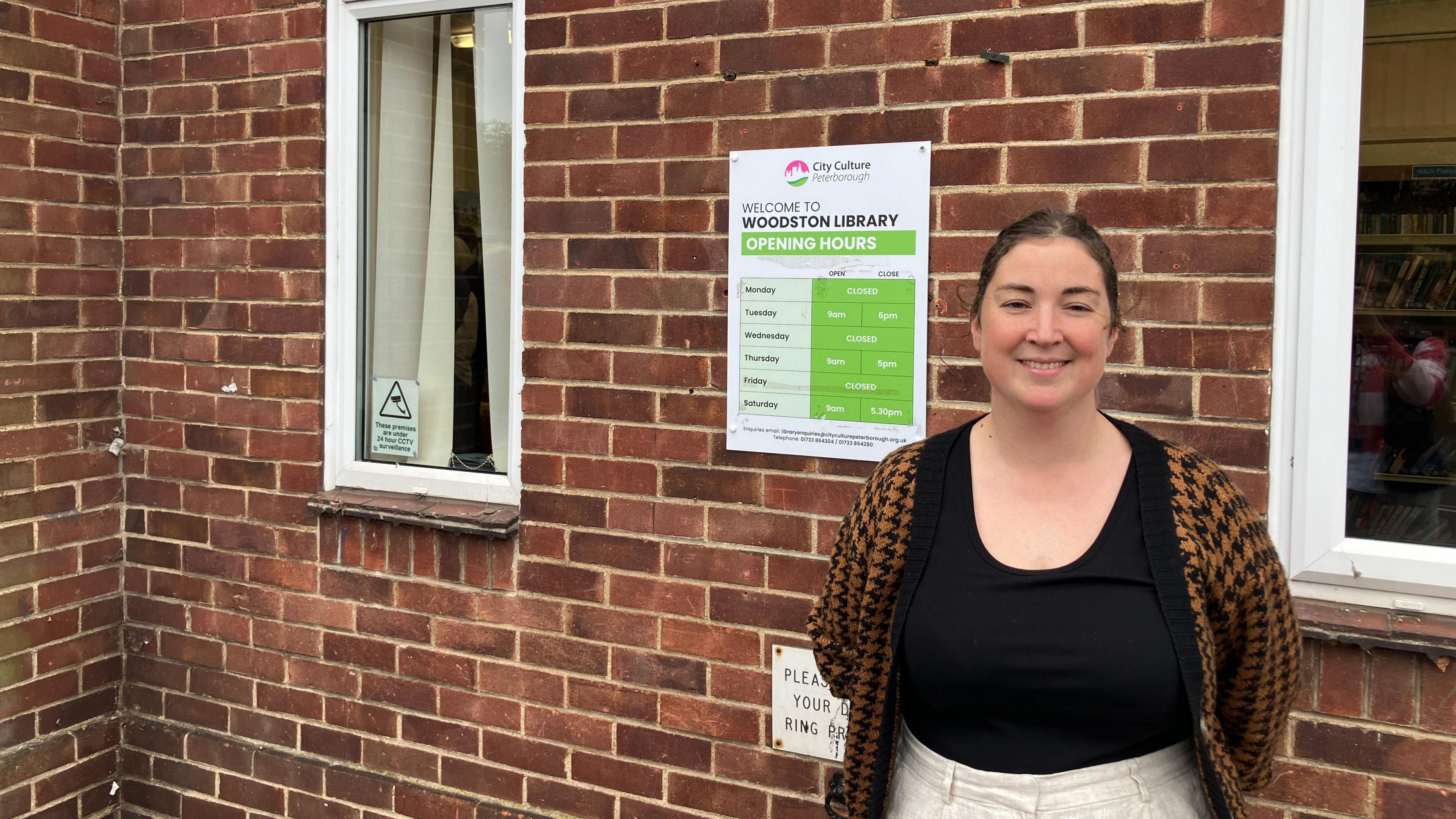 Emma wearing a black shirt and a checked cardigan standing next to the sign welcoming the public to Woodston Library