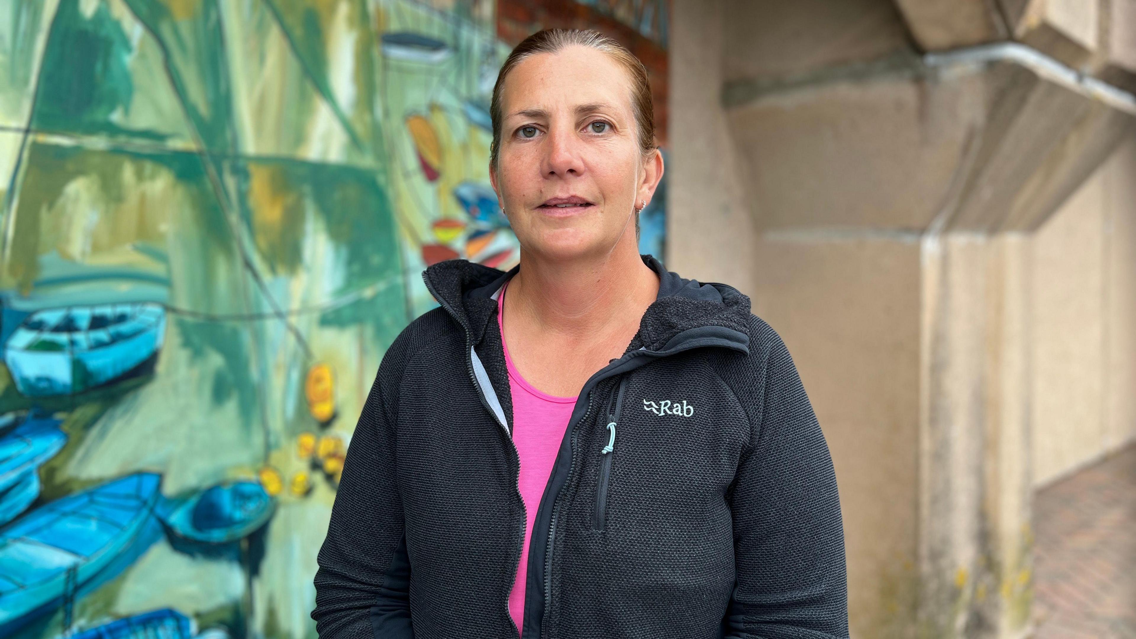 Ceri looks at the camera. She has a black fleece on over a pink running top. She's stood next to art work of boats which has been painted onto a concrete wall behind her. Ceri has brown and red hair which has been tied back.