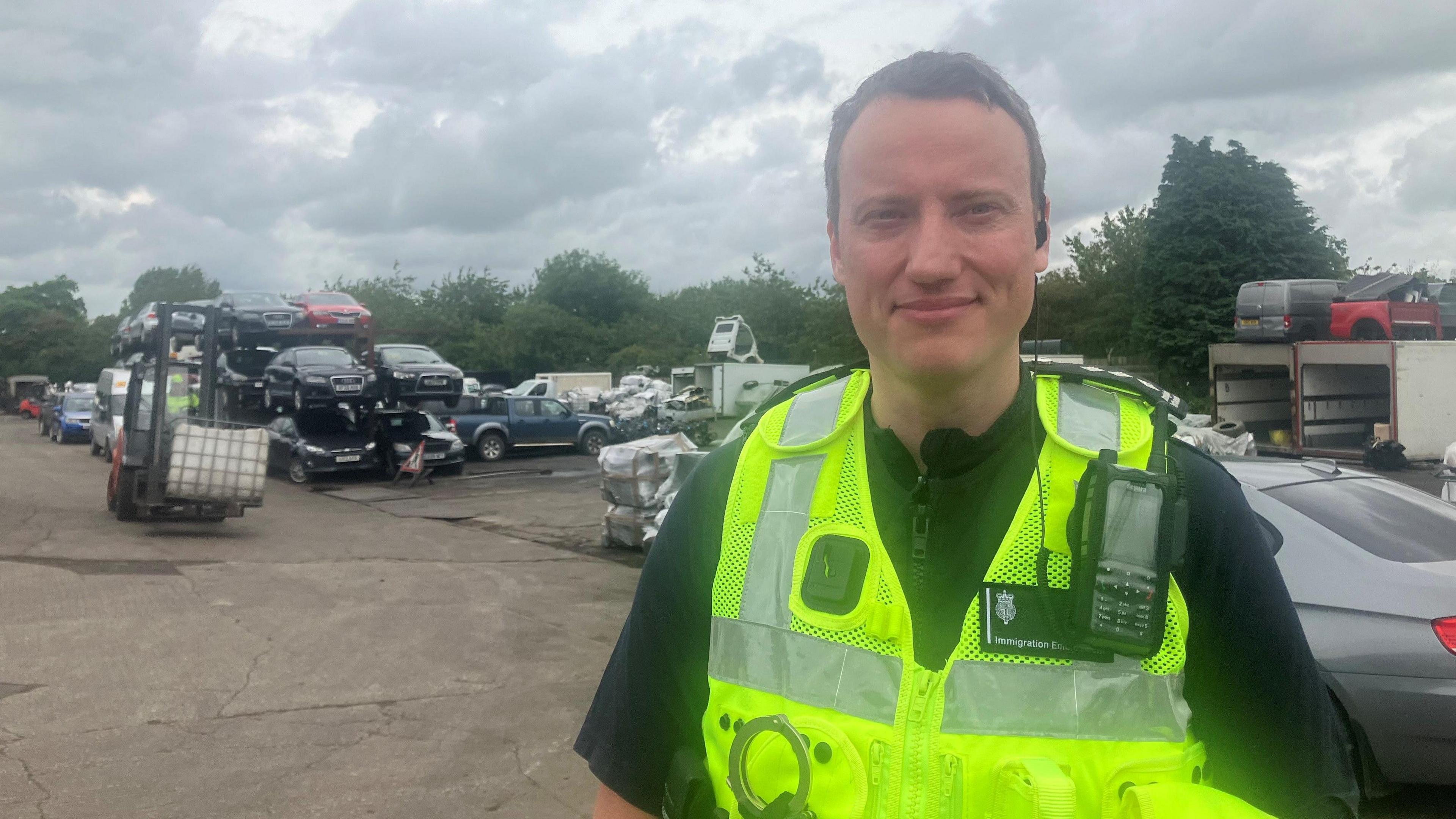 Charles Devereaux in the car scrapyard. He is wearing a high vis jacket and black top.