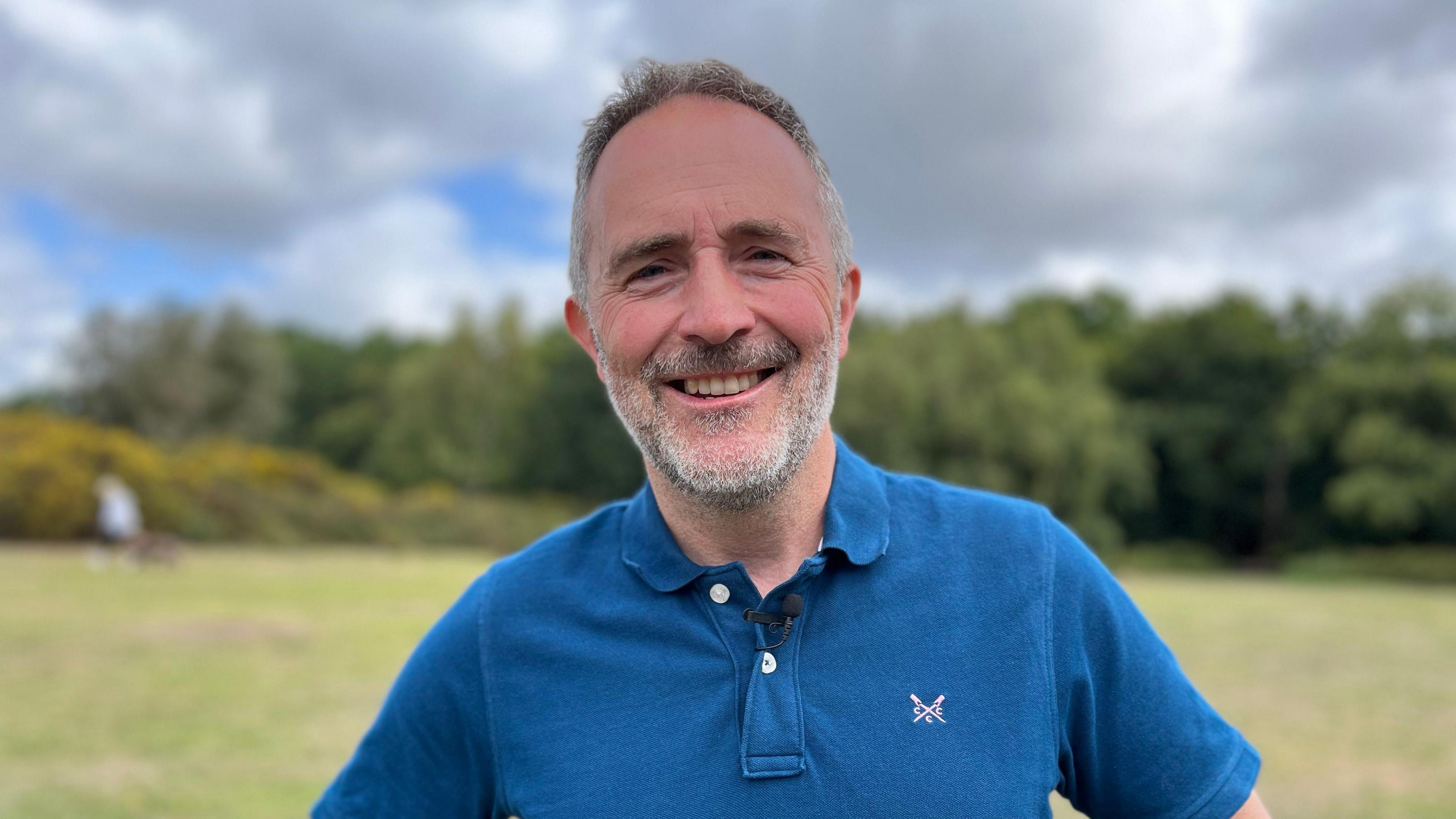 Councillor Richard Parkin stands in Sutton Park, you can see trees behind him and a cloudy sky. He is wearing a blue polo shirt and is smiling at the camera. He has grey hair and a beard.