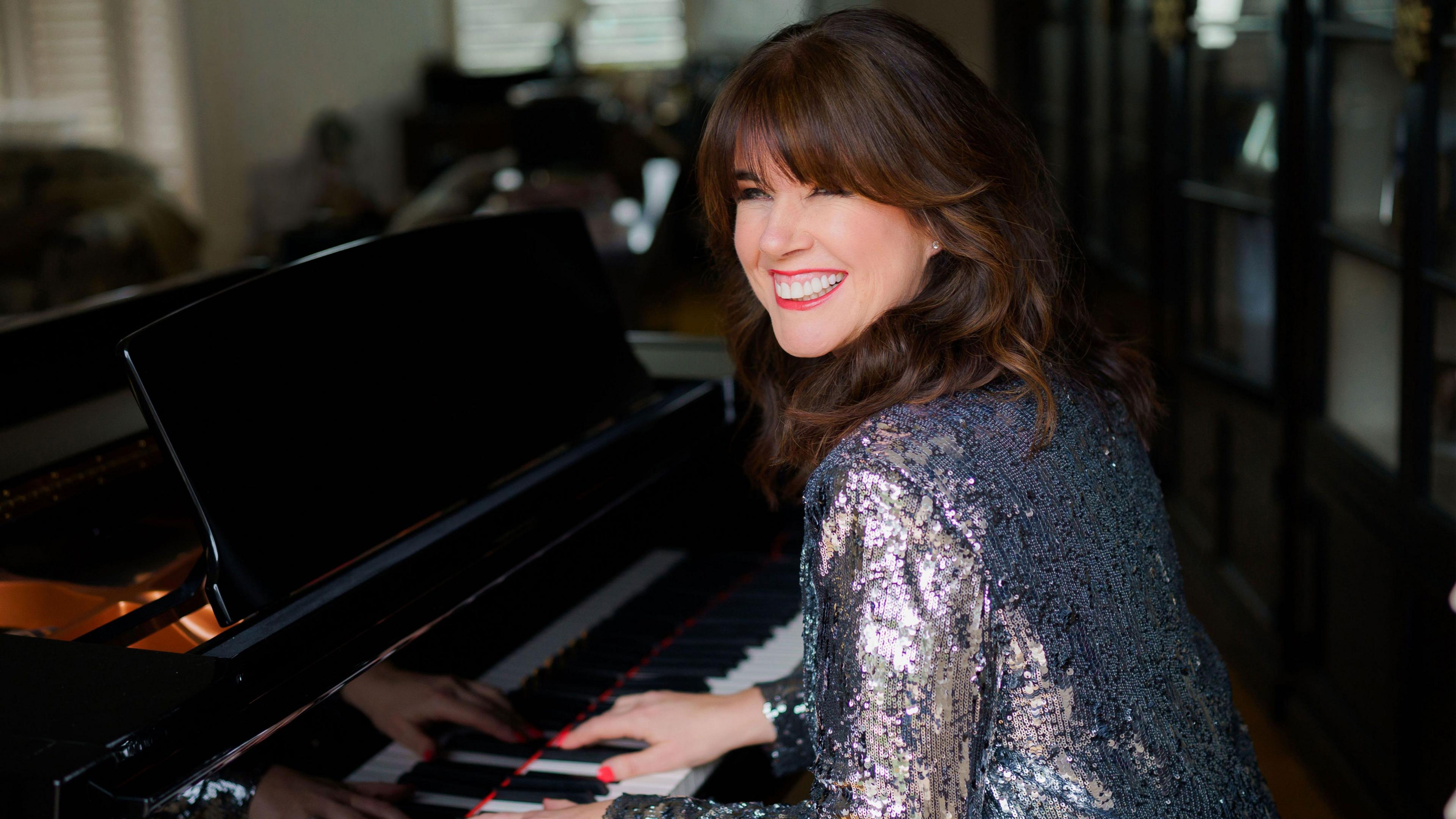 Caroline Redman Lusher sat playing a piano. She is smiling and wearing a sparkly top. 