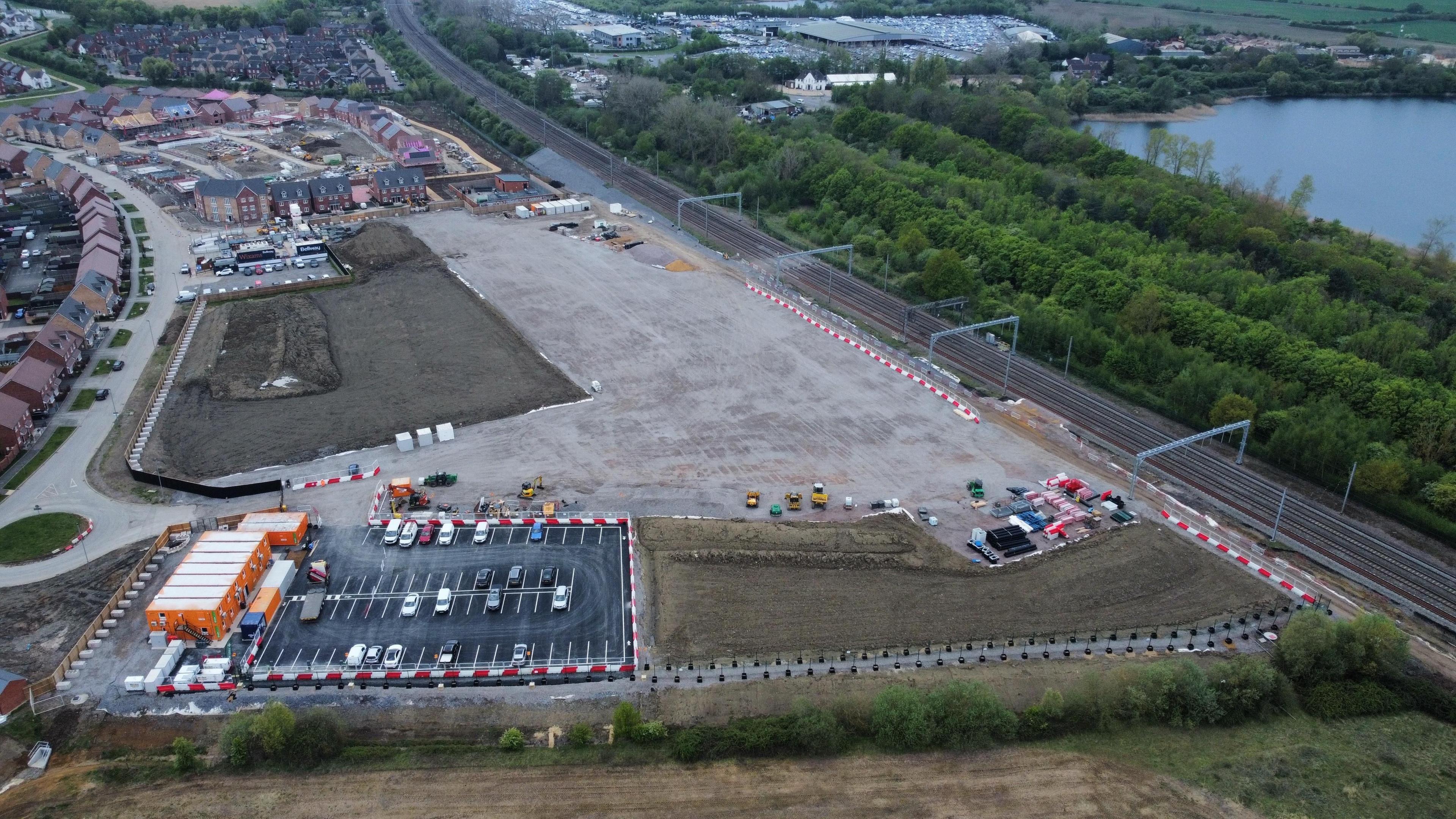 Aerial view of a construction site with scattered equipment and vehicles, nearby are residential houses and a railway track.