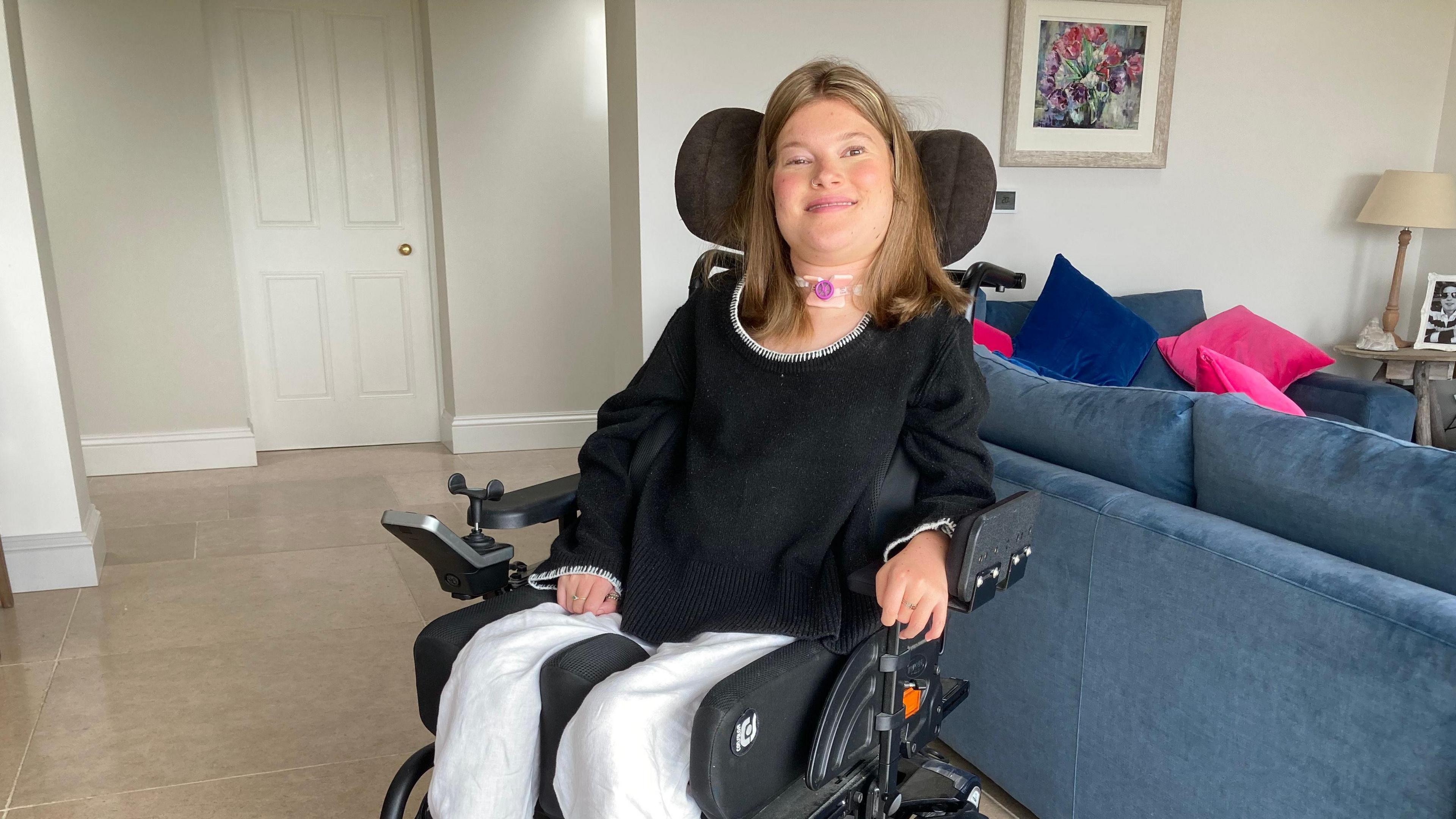 A woman in her 20s sitting in a living room in a wheelchair. She is smiling at the camera and wearing a black top. Two blue sofas are behind her.