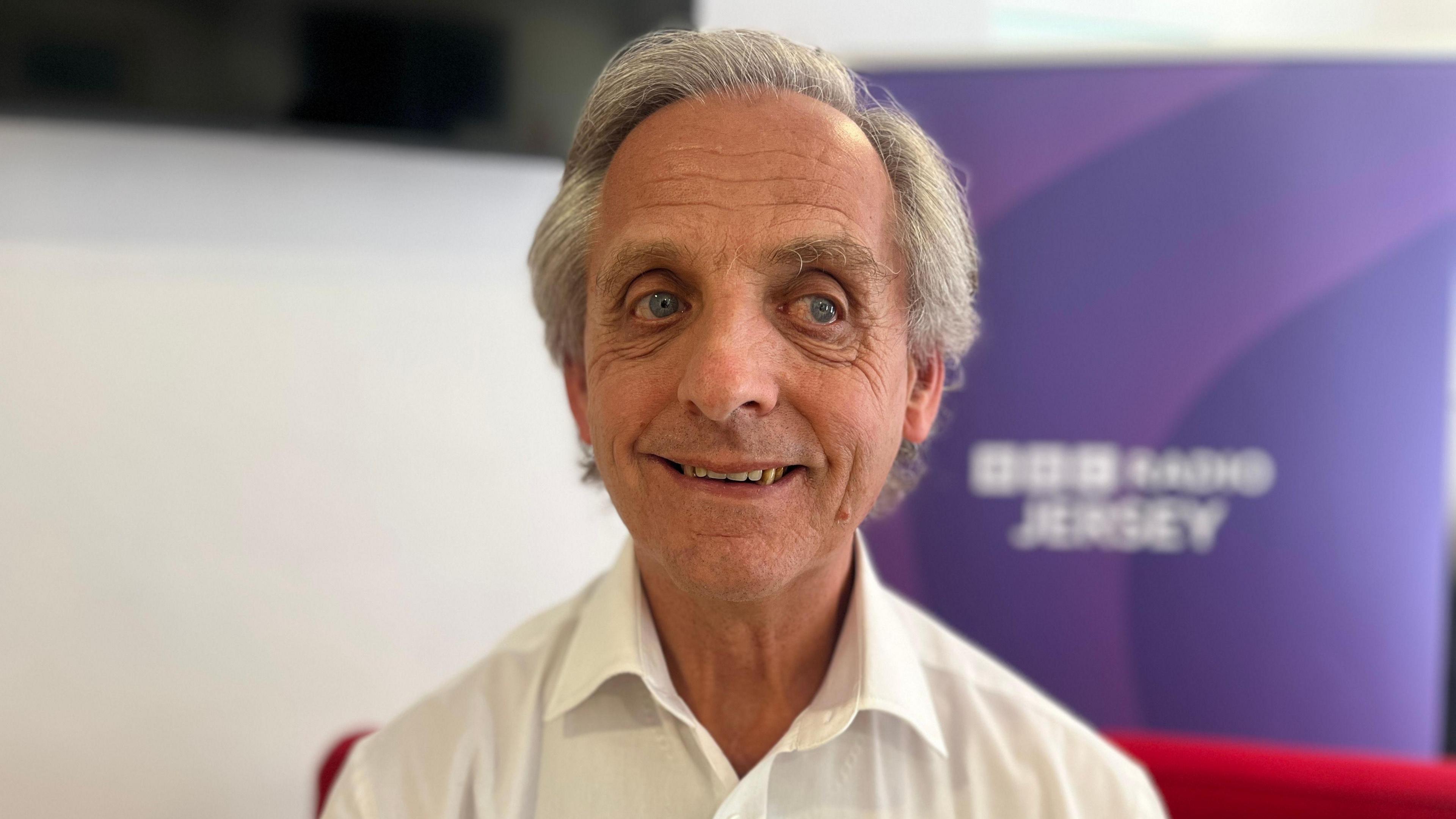 Nicholas Ozouf smiles while sat on a red sofa in front of a purple BBC Radio Jersey banner