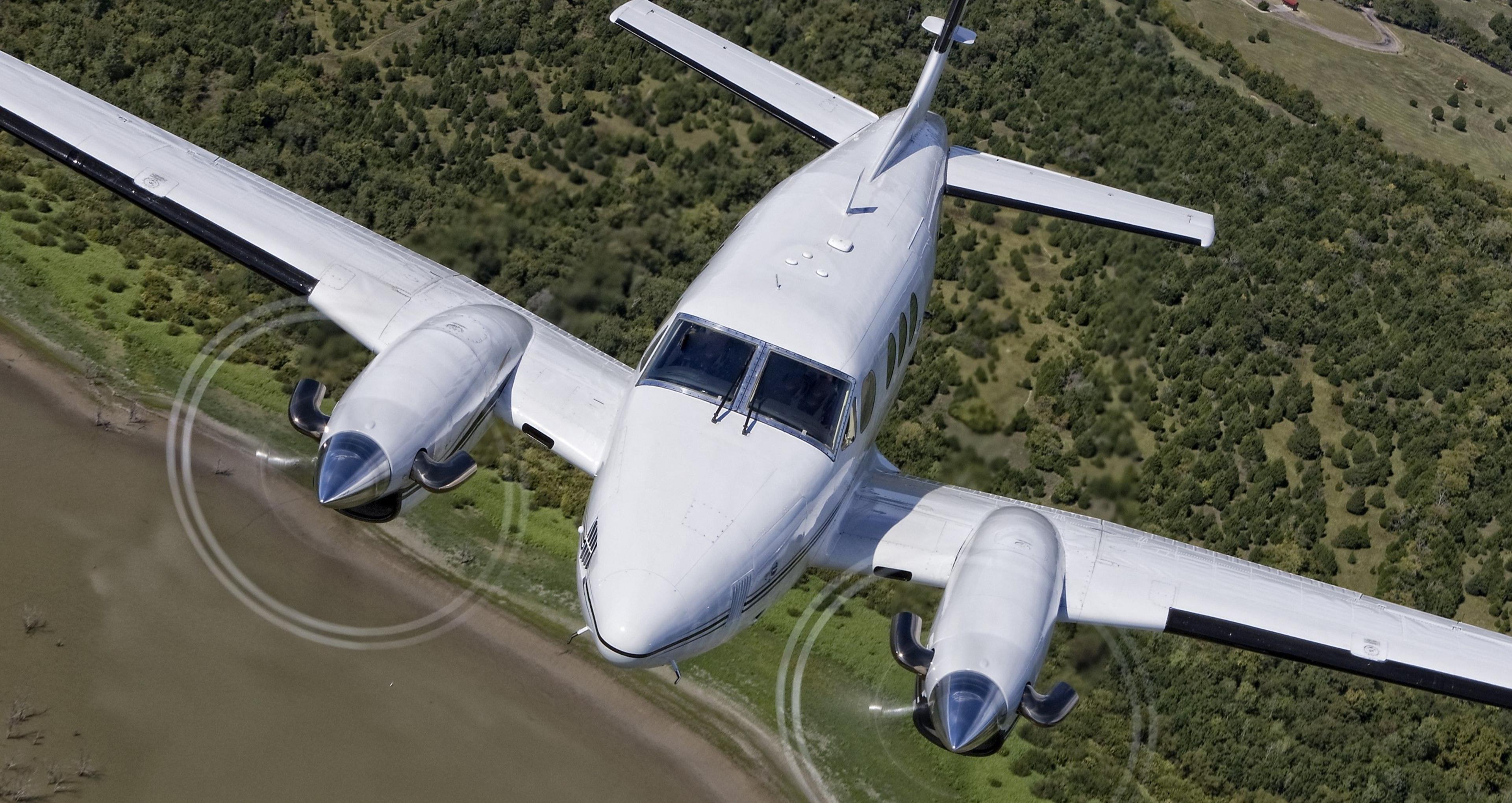 A blue and white plane.  Its propellers are moving. It is ascending. There is greenery underneath.