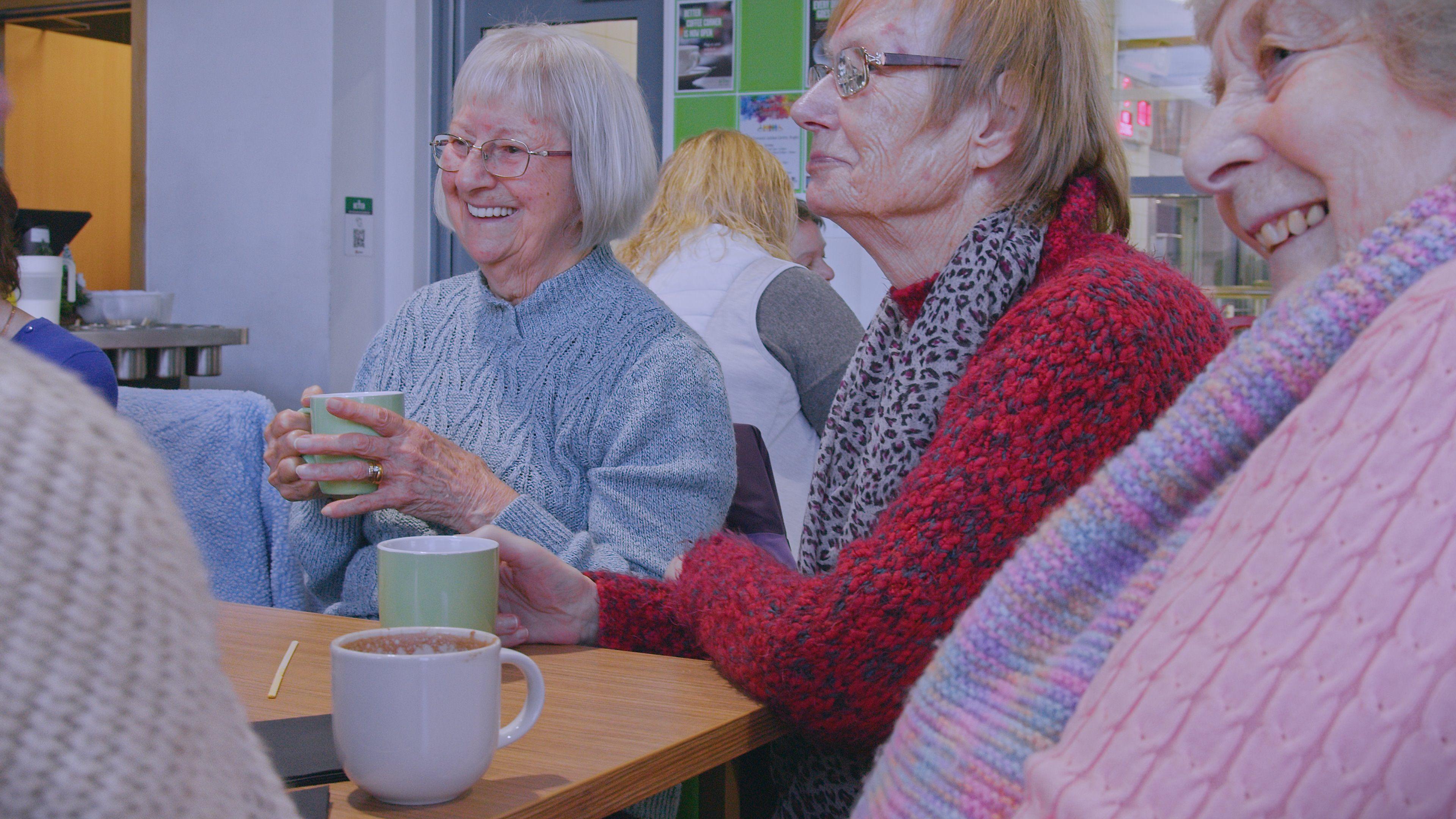 A group of women sitting around a table and drinking hot beverages. One woman is holding a green cup, with a wedding ring on, and smiling. She is wearing a light blue jumper, and has fair hair in a bob. The woman next to her has a leopard print scarf on, a red jumper and is holding a cup in front of her. The other woman has a pink scarf and jumper on and is sitting back. 