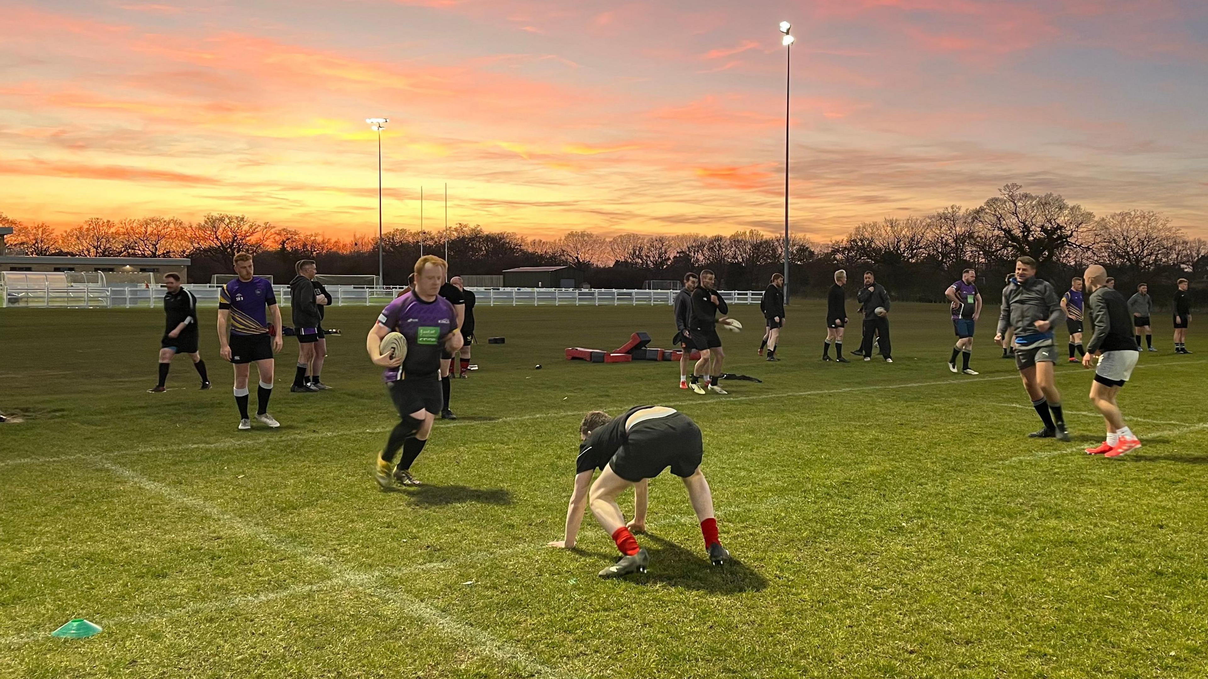 Colchester Kings players are taking part in tackling practice during a training session on a sports field. There are about 15 people in the picture in T-shirts and shorts. There are floodlights switched on and the sun is setting.