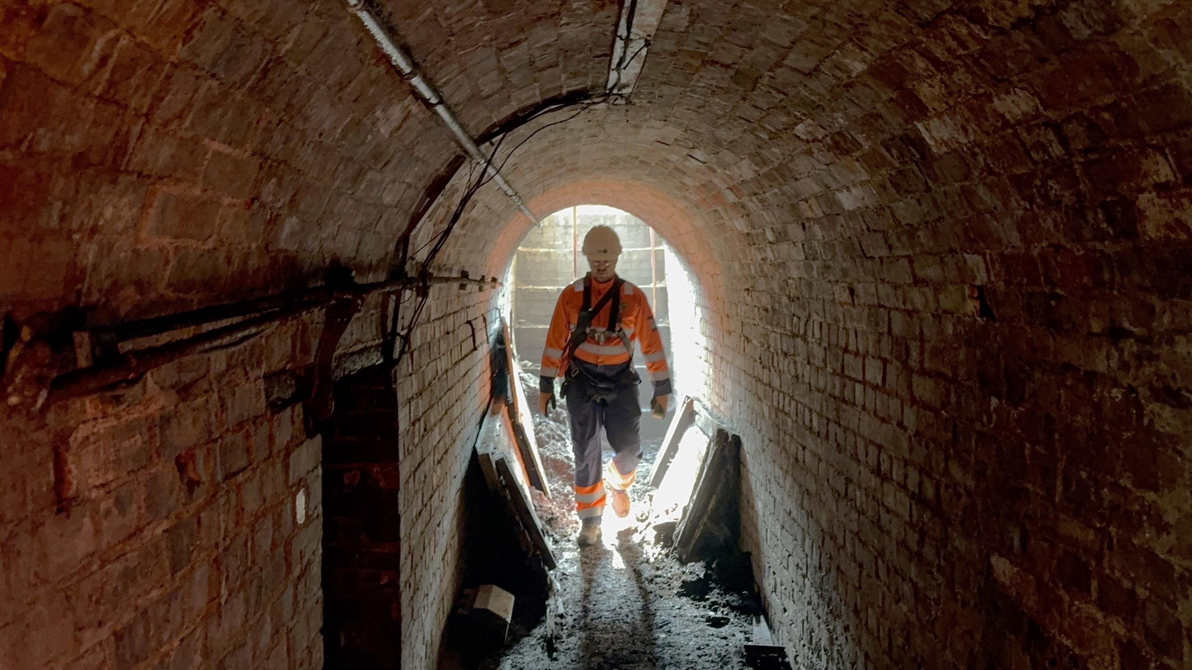 A man wearing an orange hi-vis suit, dark coloured trousers and a hard helmet. He is walking through a dark brick tunnel towards the camera, and is slightly silhouetted from light behind him. The tunnel floor is filled with debris, dirt and dust. There are wires fixed to the walls of the tunnel.