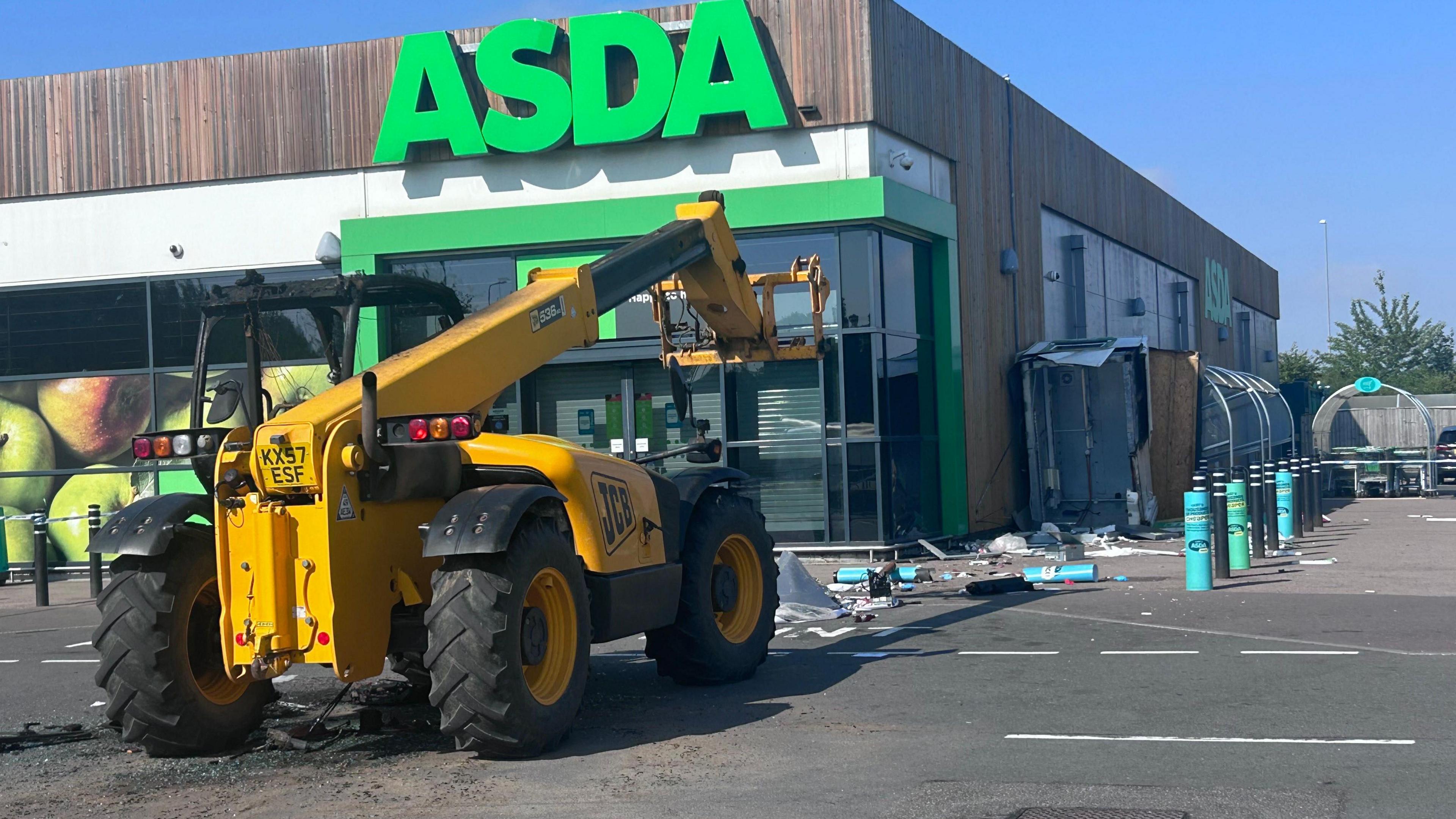 Telehandler outside a branch of Asda - a large green-coloured Asda logo is on the store front. Debris from smashed bollards is strewn in front of the telehandler. 