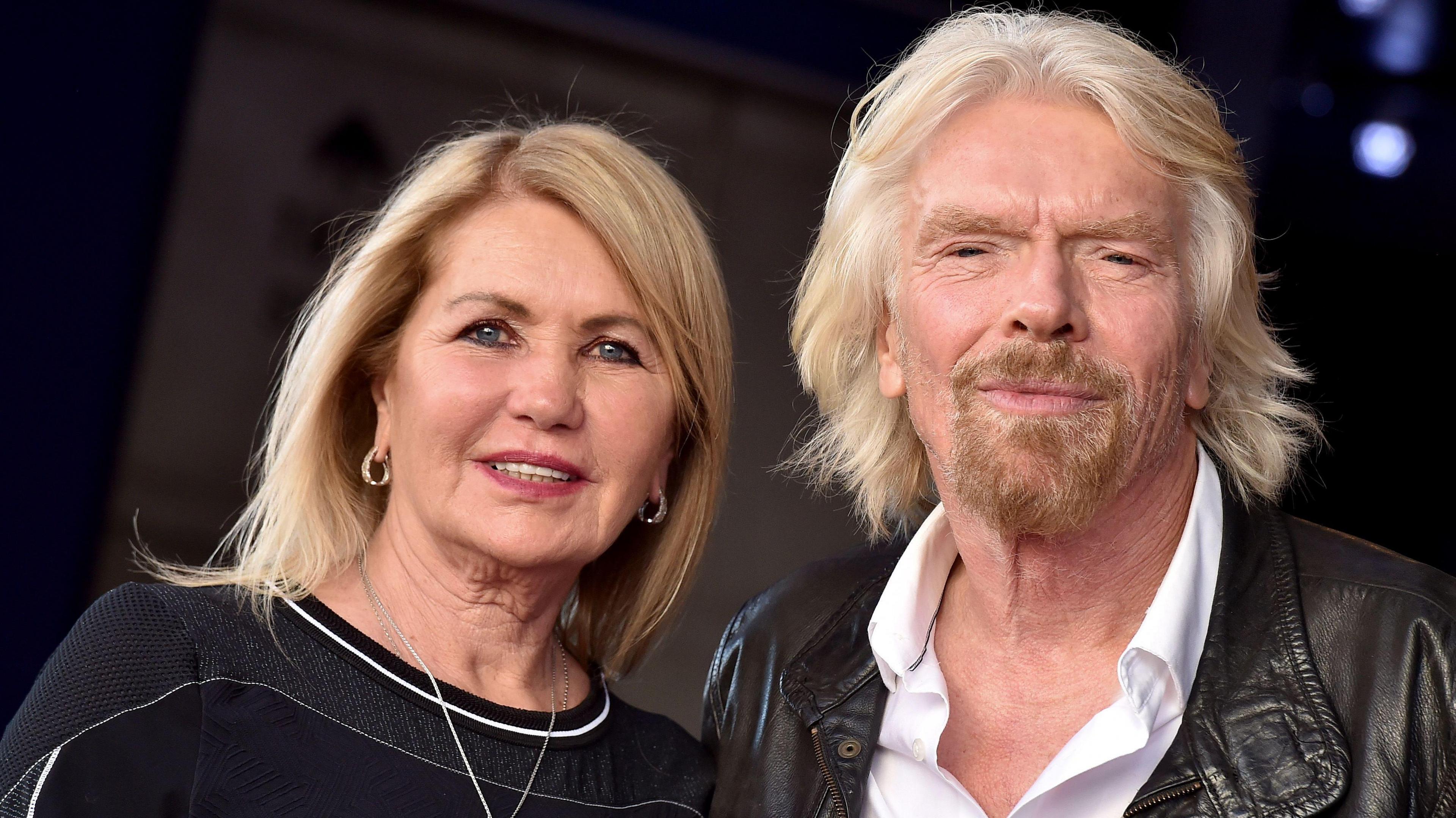 Joan Templeman and Sir Richard Branson look towards the camera. She has long blonde hair and a dark top. Branson has light long hair, a beard, white shirt and brown leather jacket. 
