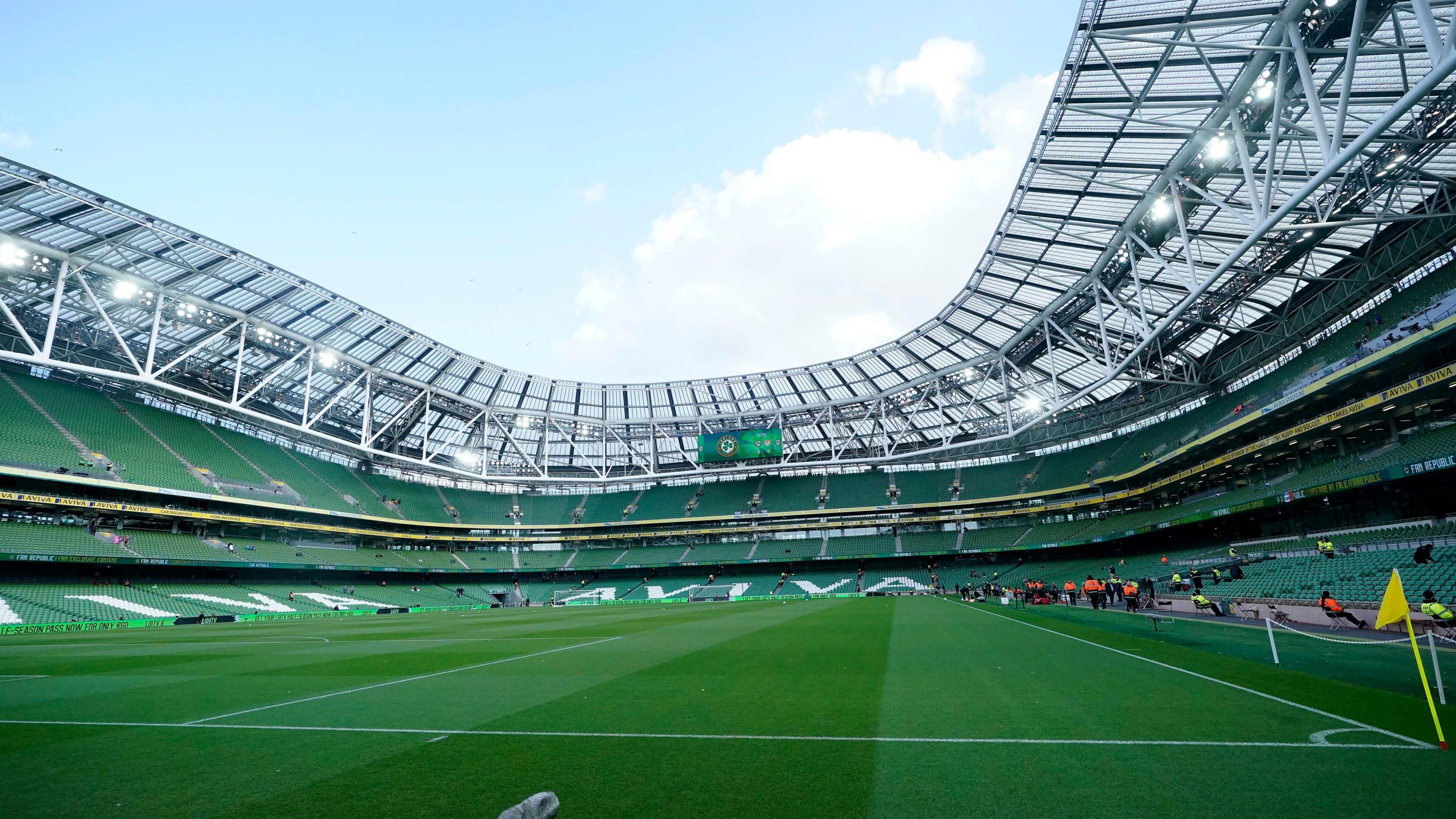 An empty Aviva Stadium in Dublin, shown when no concerts or sporting events are taking place. Three quarters the stadium and three tiers of green seats can be seen surrounding a lush football pitch. 