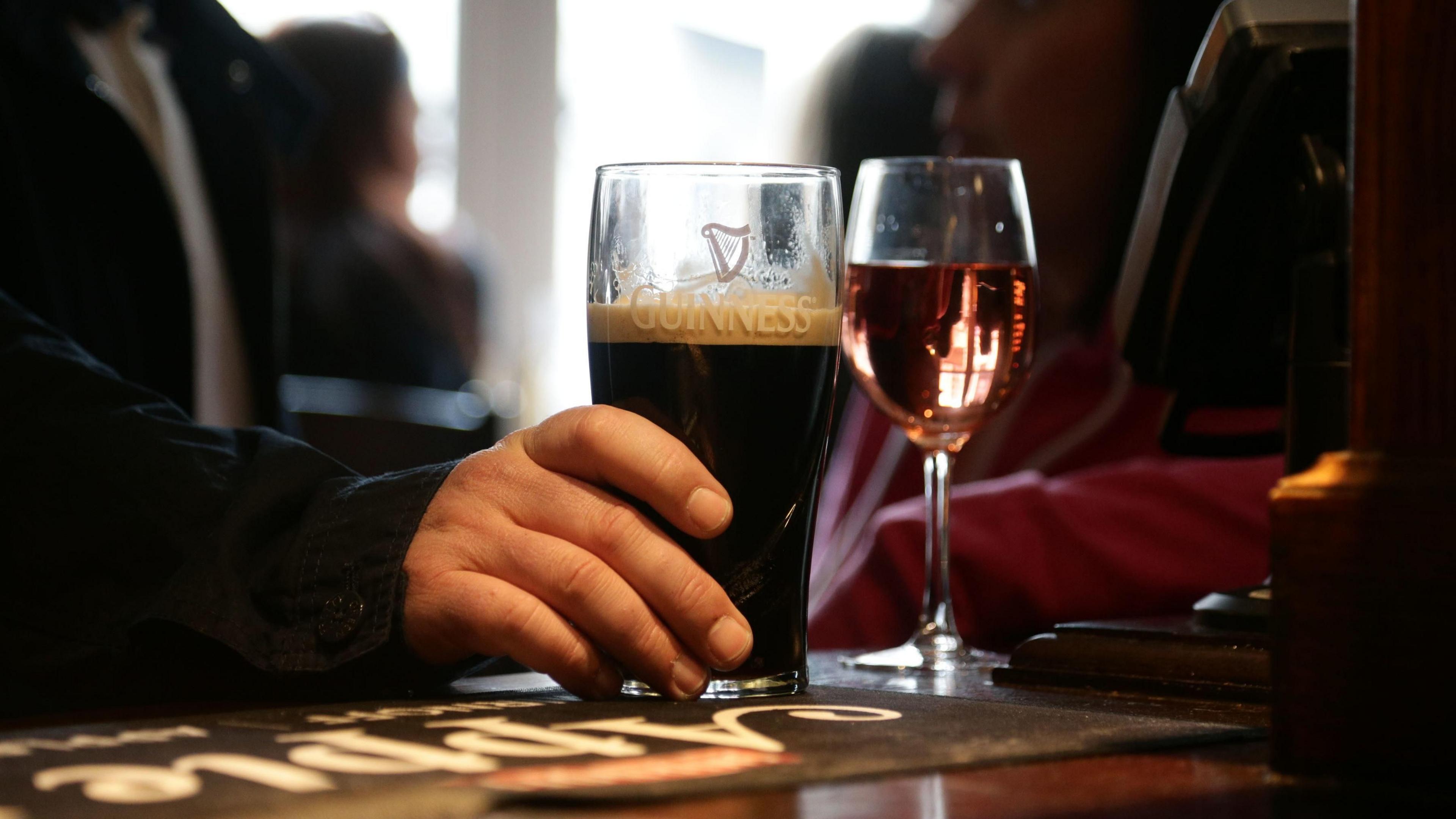 A pint of Guinness and glass of rosé sit on the bar of a pub. A man has his hand on the pint glass.