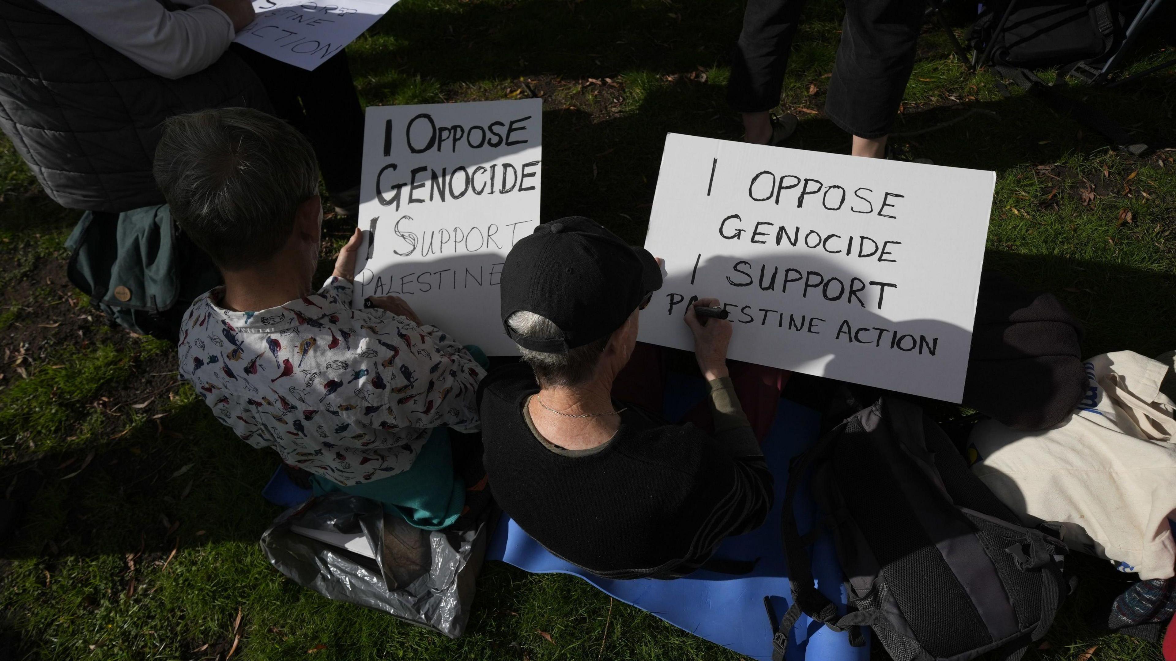 Two protesters sit on the grass writing placards 'I oppose genocide, I support Palestine Action'