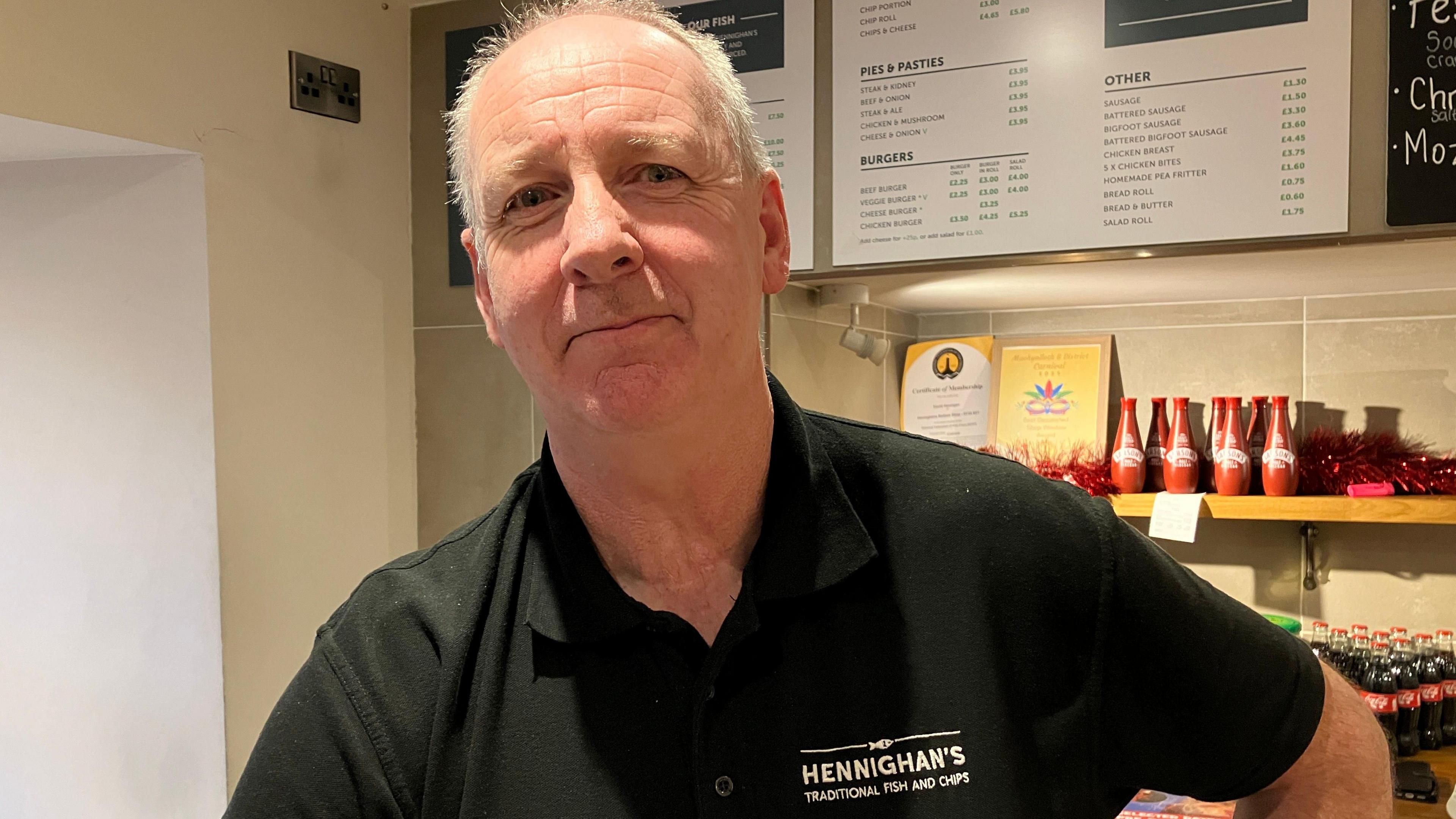 A man is wearing a black polo and is working behind the counter of  fish and chip restaurant. Behind him are boards with costs, and various items such as bottles of vinegar.