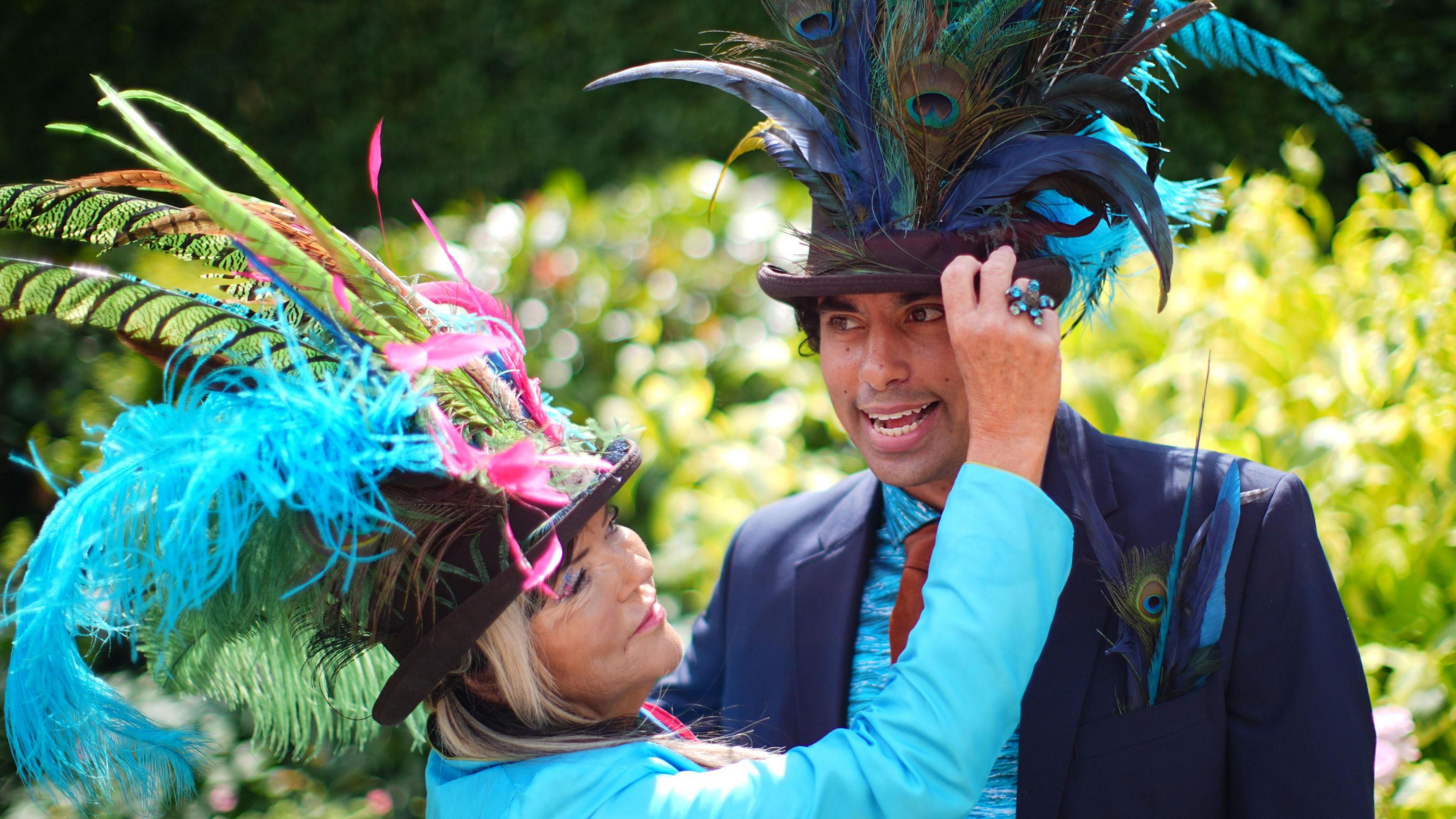 A man wearing a dark suit and a top hat covered in peacock feathers is tended to by a woman wearing another elaborate hat with feathers coming out of it, who is also wearing a light blue jacket, at Ascot Racecourse.