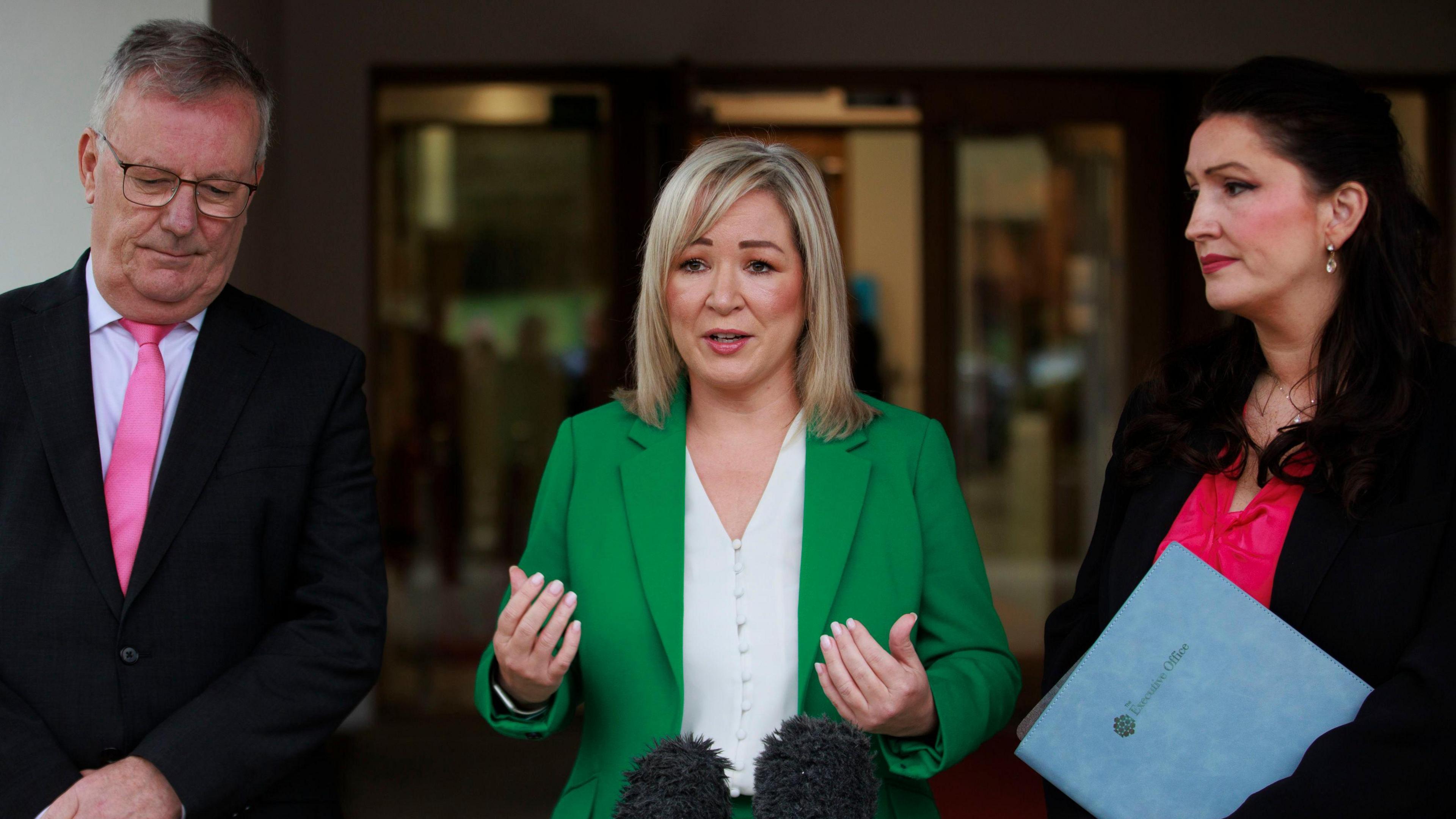 Mike Nesbitt, Michelle O'Neill and Emma Little-Pengelly standing around a microphone. Mike Nesbitt is wearing a black suit and pink tie with a white shirt. Michelle O'Neill is wearing a white shirt and green blazer while Emma Little-Pengelly is wearing a black blazer and pink top. She is carrying a blue folder.