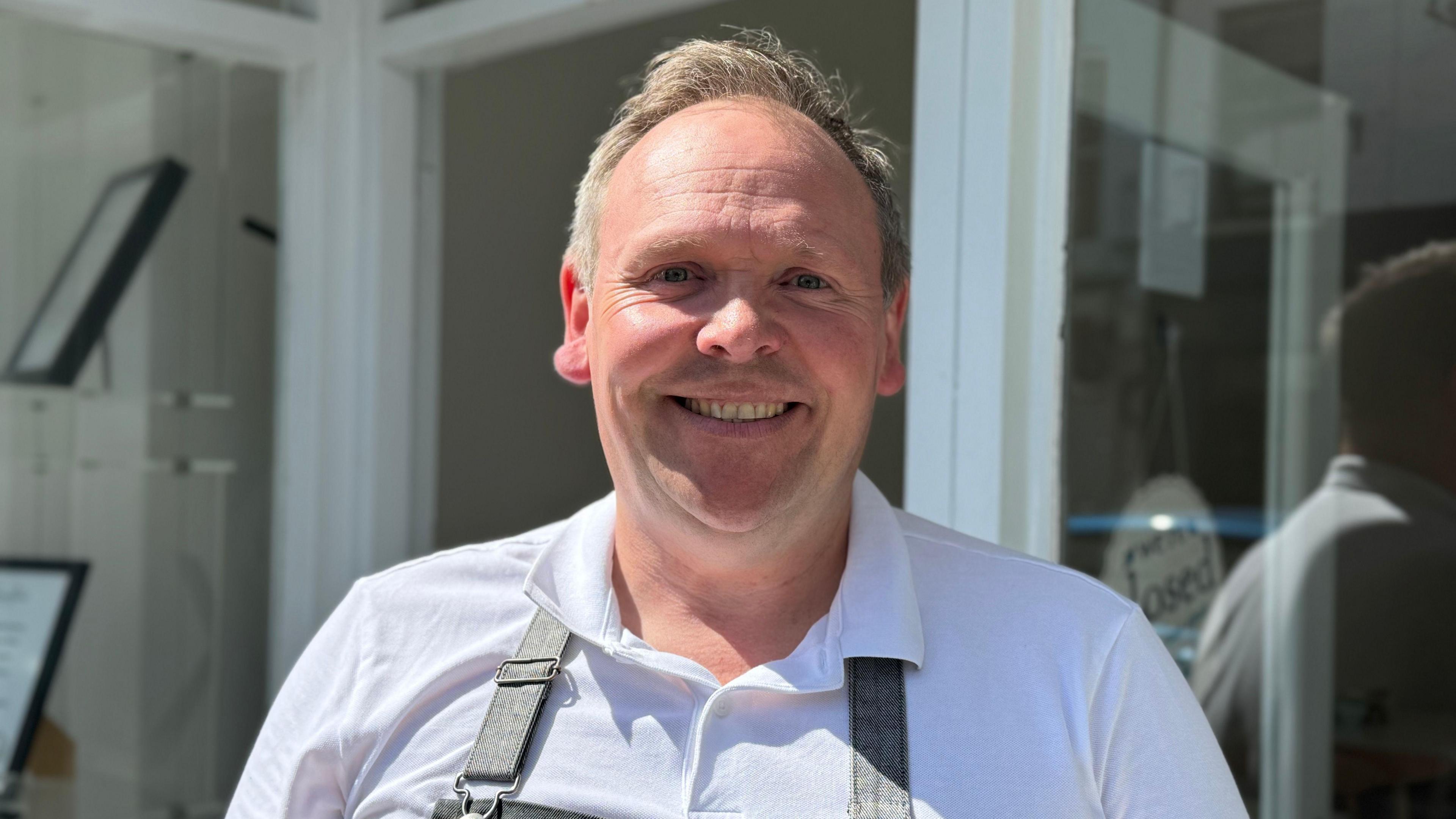 Steve Hamilton from Hamilton's Tea House looks straight at the camera. He is wearing a white top with a collar. You can see the start of an apron. The windows of the tea house can be seen behind them.