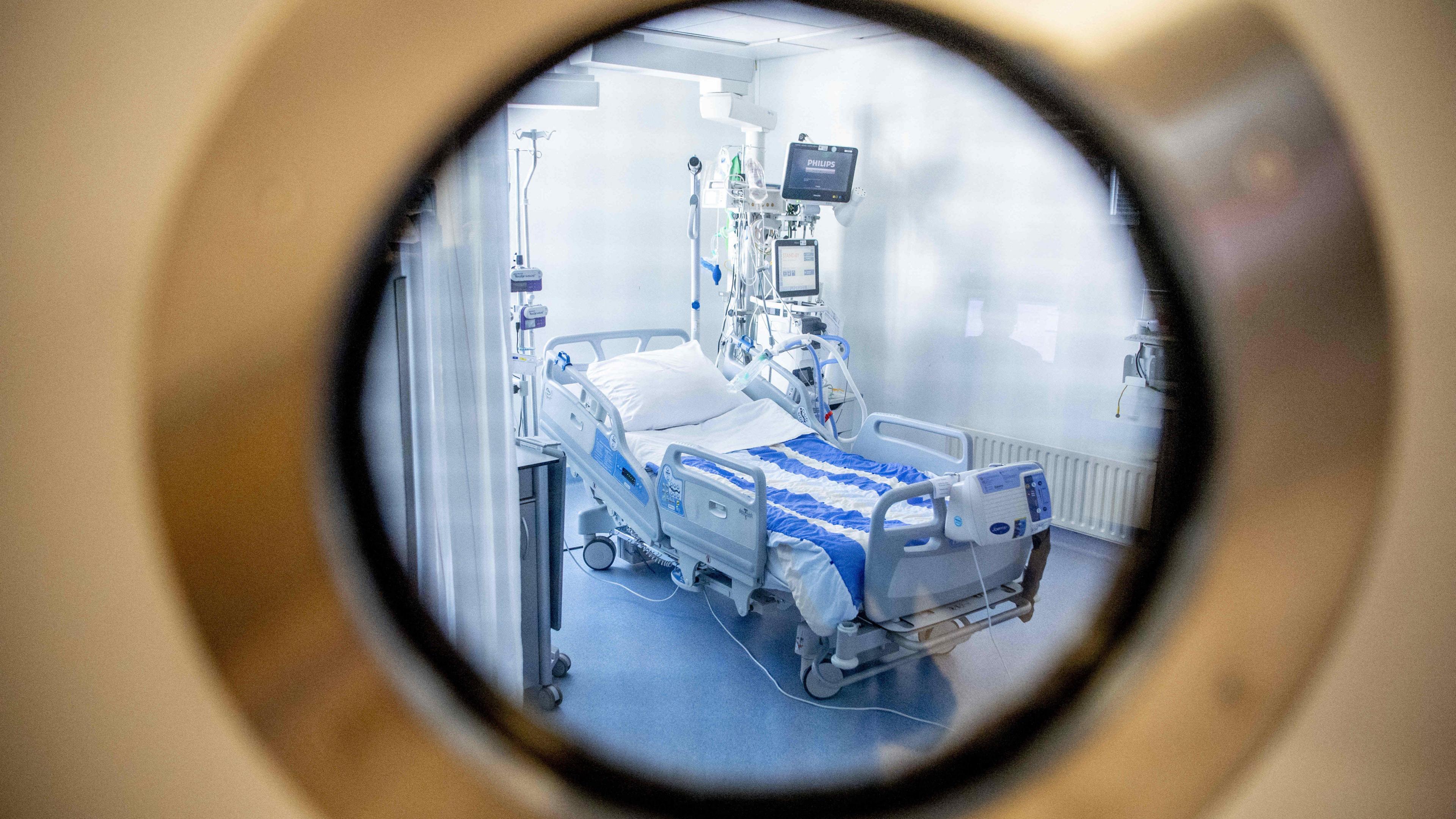 Looking through a round window into a hospital room which has a bed with wheels with a row of monitors next to it.