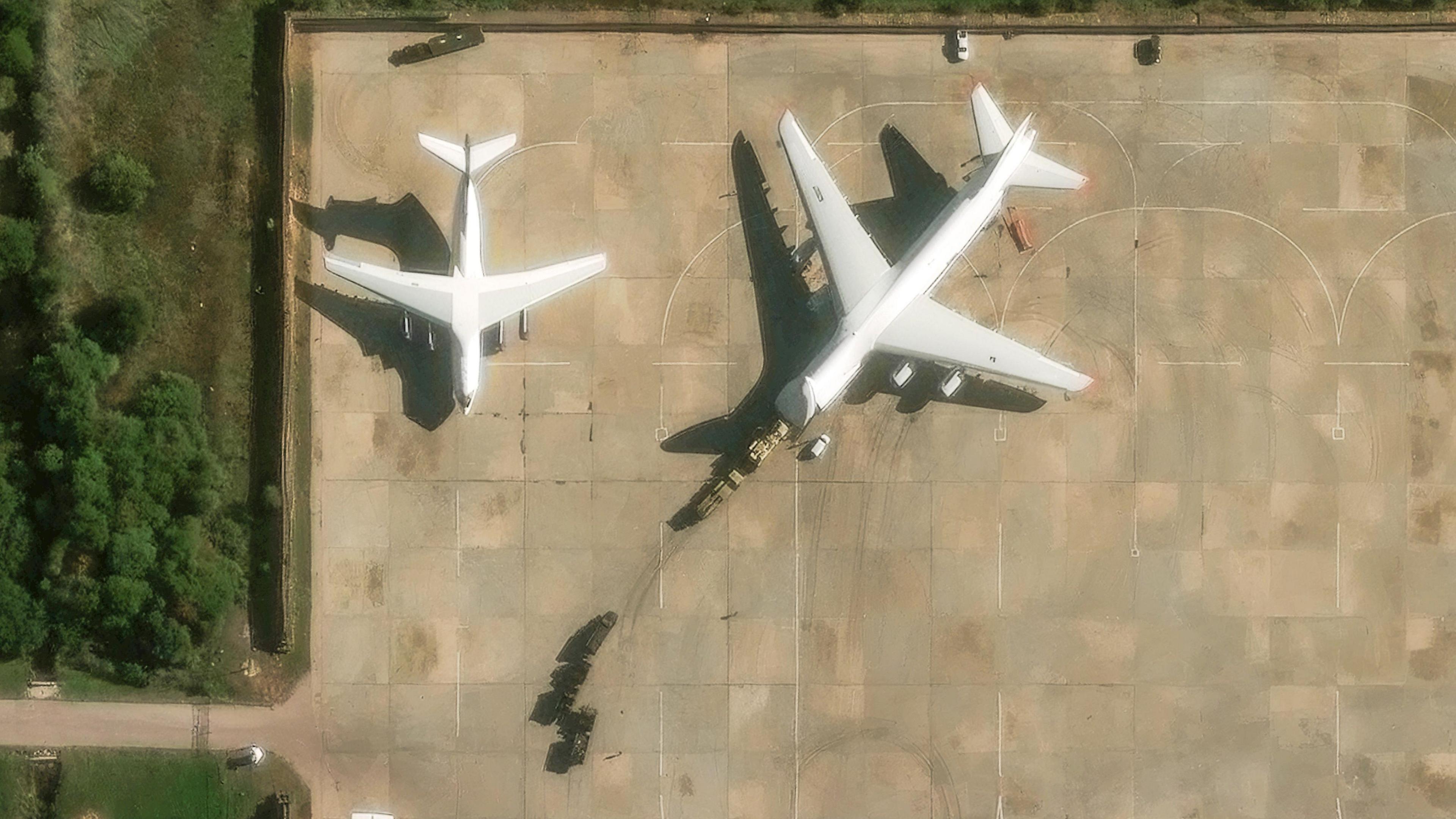 Two planes are parked on the runway at Hmeimim air force base. Military vehicles can be seen driving onto one of the planes, with further vehicles parked behind. 