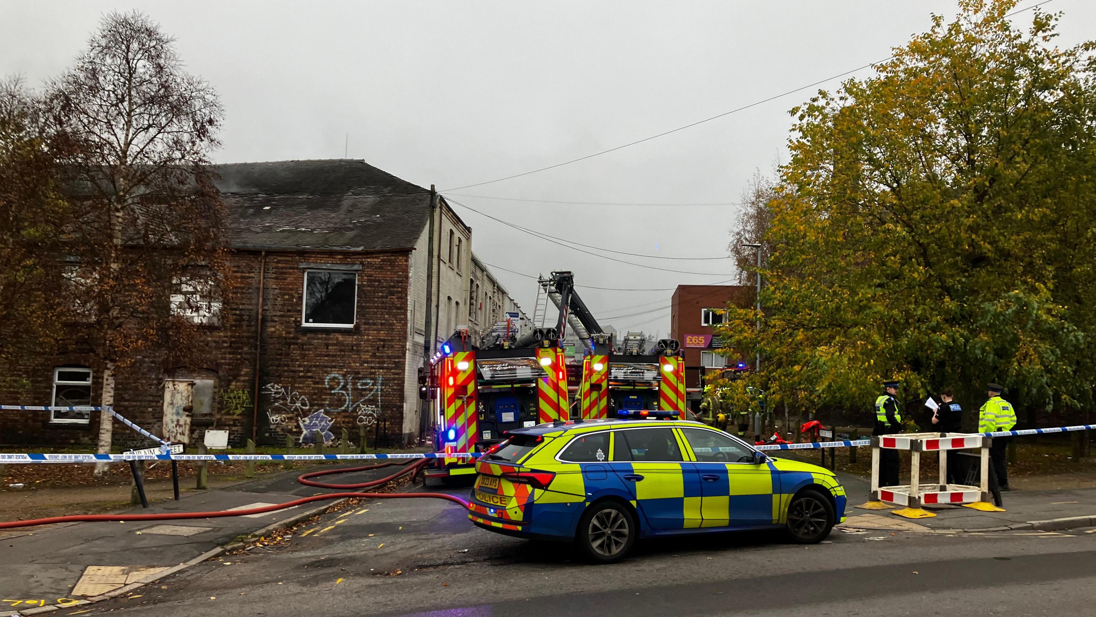 Firefighters and police officers work behind a cordon with police tape blocking off the area where two fire engines are situated. Several police officers can be seen to the right next to a large tree.